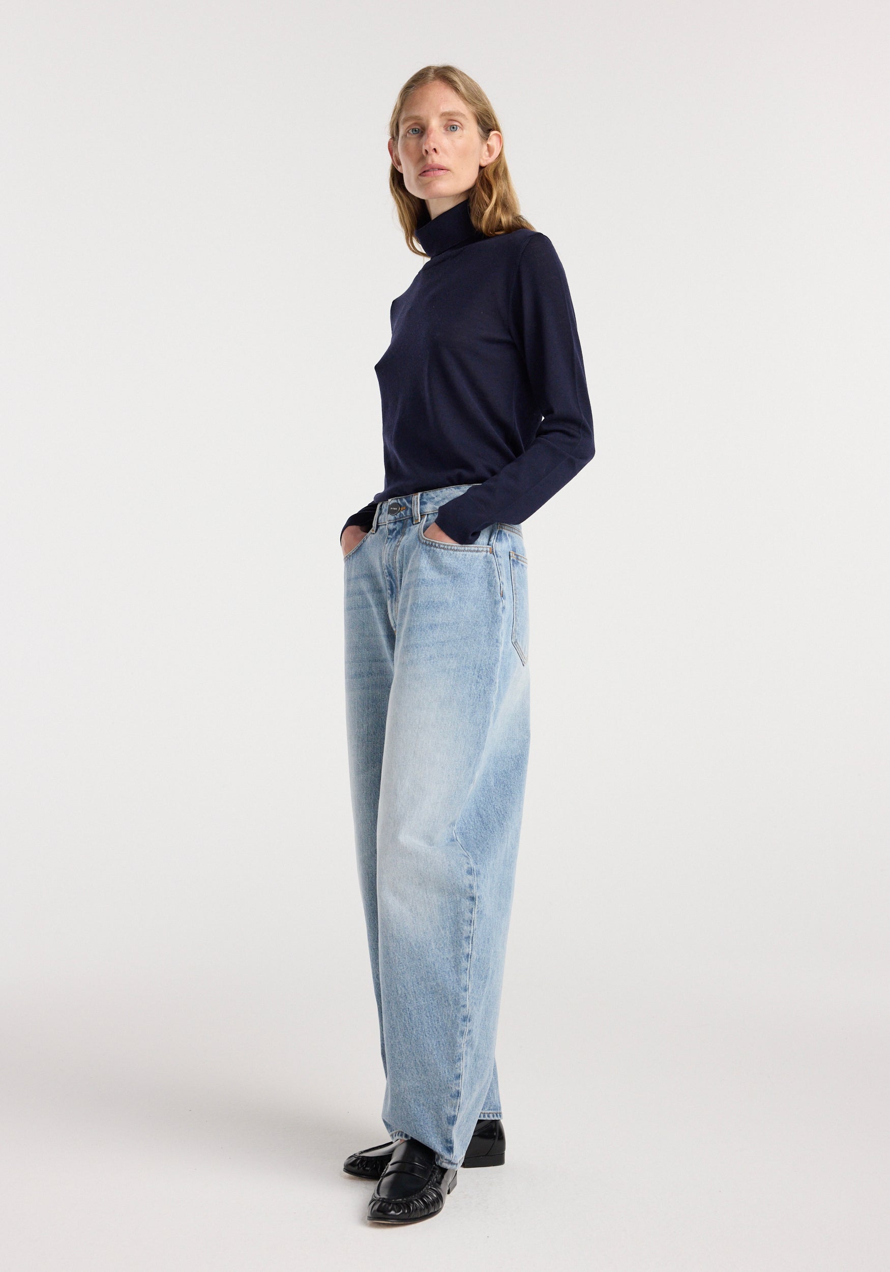 Woman wearing Fransje - Sculptural denim trousers | classic blue with a high waist and wide-leg fit paired with a dark navy long-sleeve turtleneck sweater and black shoes.