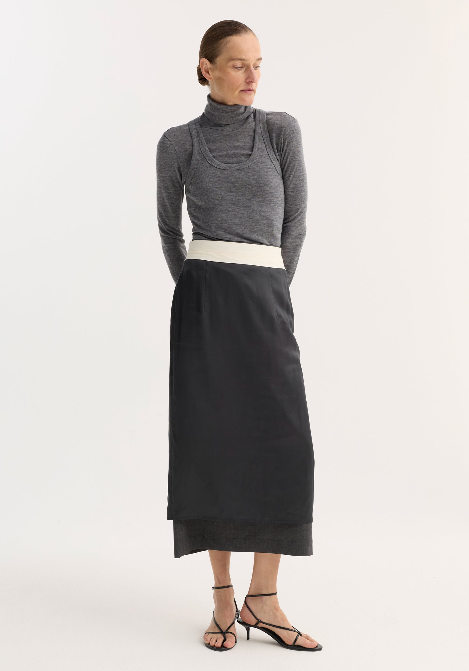 Model in fitted Double-layer wool top, mid grey melange, featuring a turtleneck and tank combo, paired with a high-waisted black skirt and sandals.