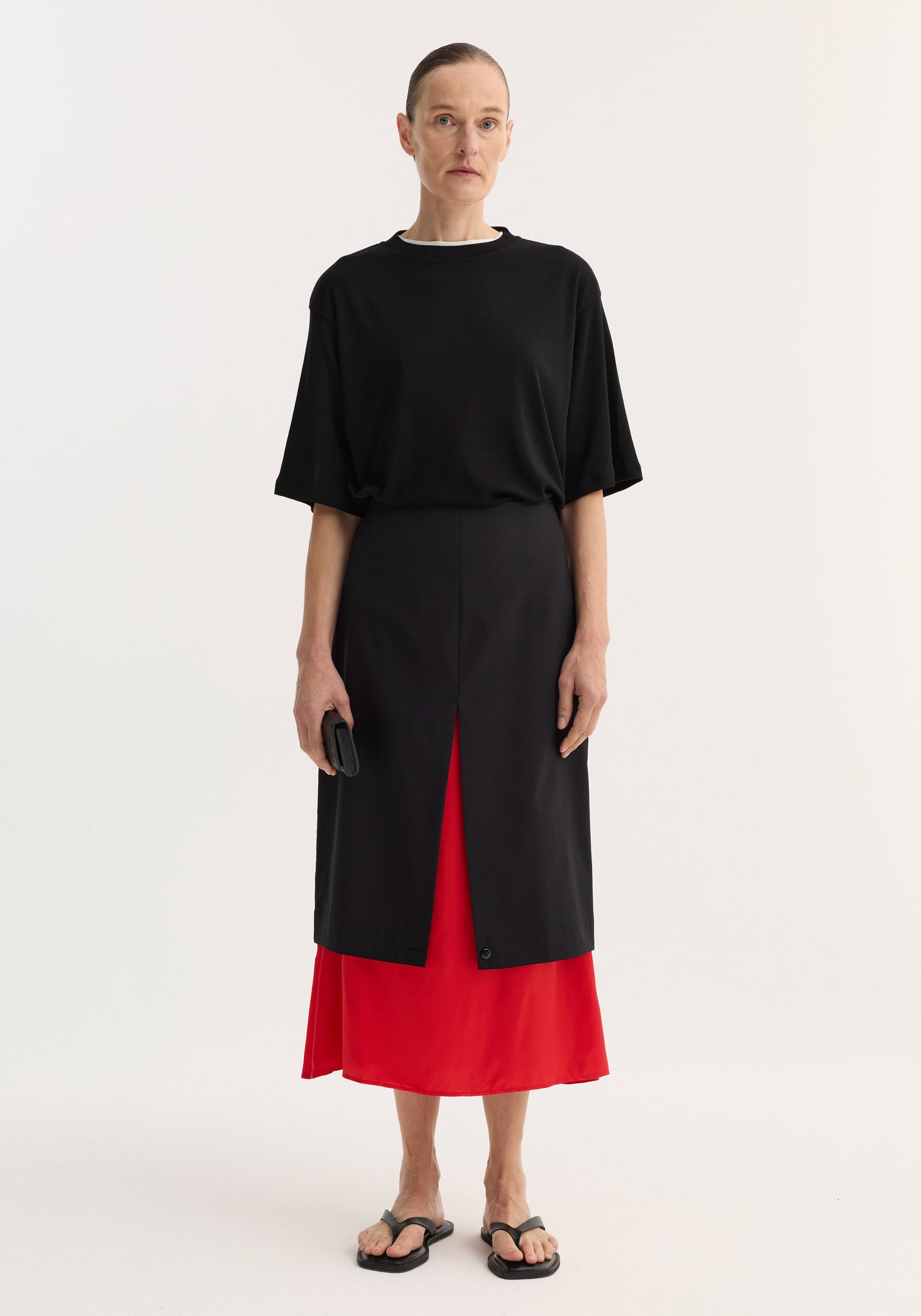 Model wearing the Contrast upside-down skirt in black, featuring a front slit with a red underlayer, paired with an oversized T-shirt and black sandals.