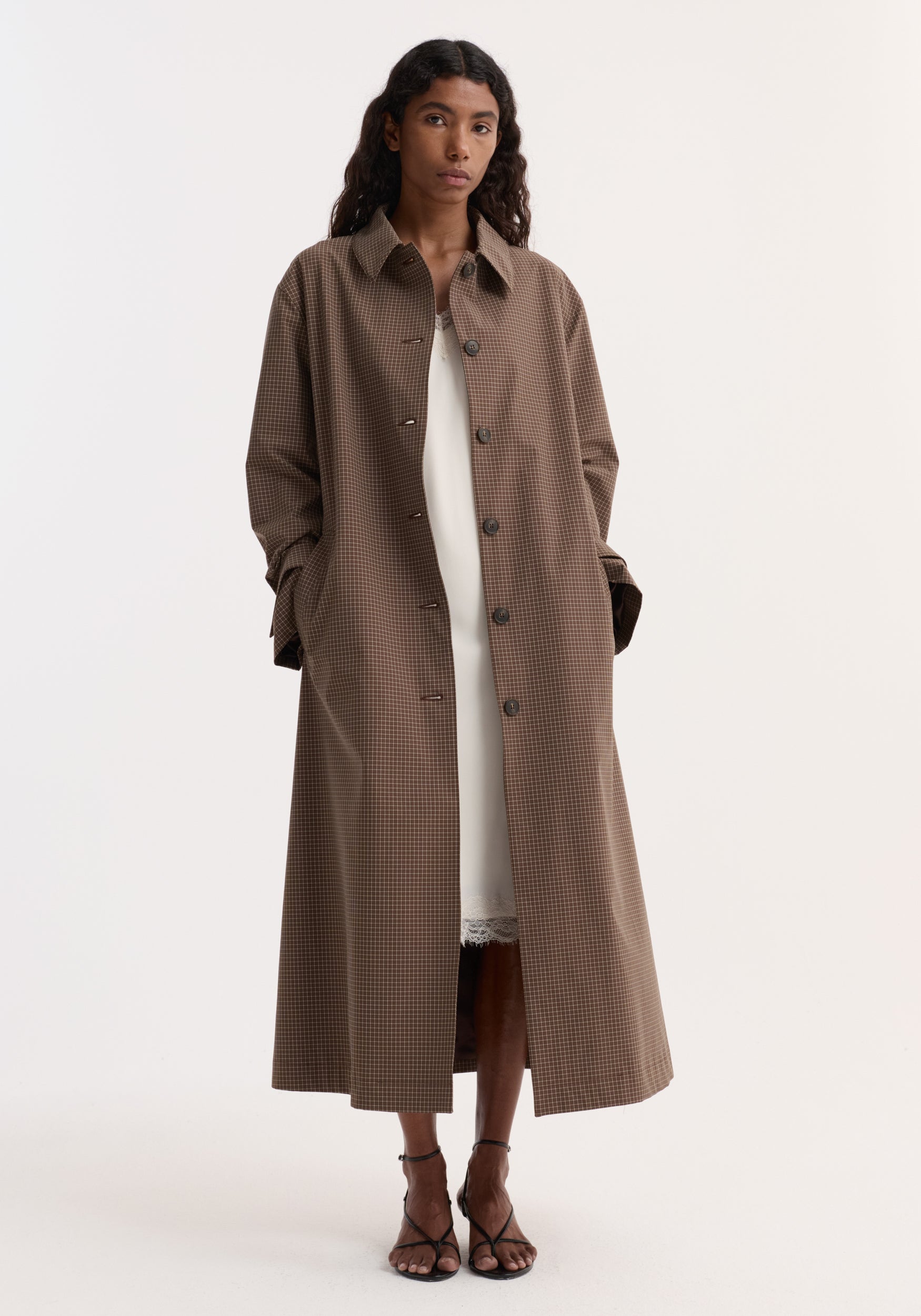 Model wearing a long Technical check belted coat | brown multi check with a button-up front, collar, and pockets, paired with a white dress and black strappy heels.