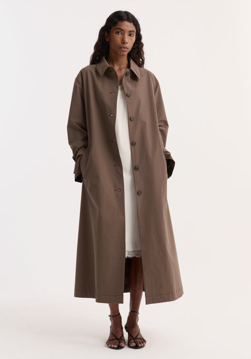 Model in Technical check belted coat, brown multi check, with a tie belt, structured collar, and cuff straps, over a white slip dress.
