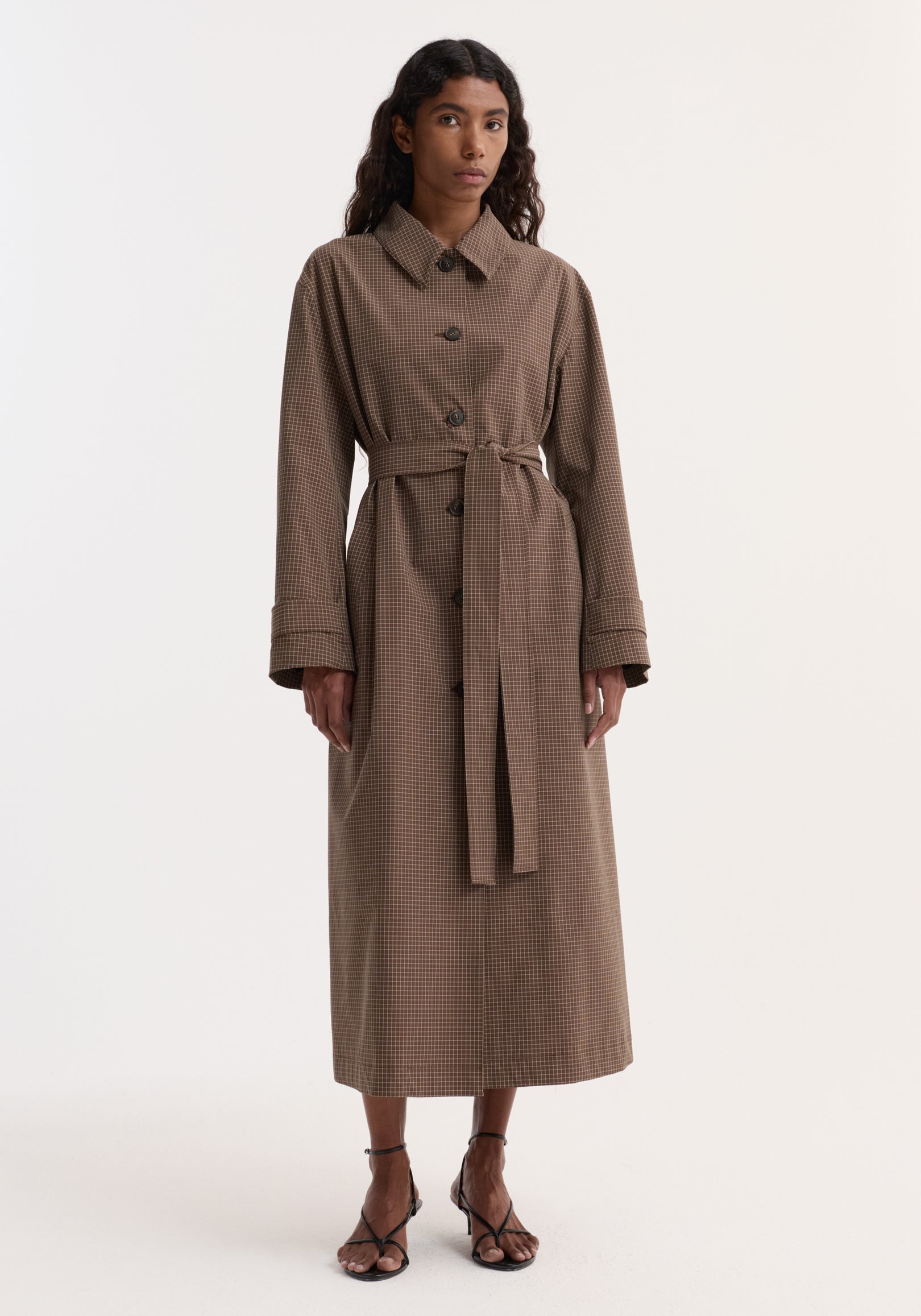 Woman wearing a long Technical check belted coat | brown multi check with button closure, wide collar, cuffed sleeves, and a matching belt tied at the waist, paired with open-toe heels.