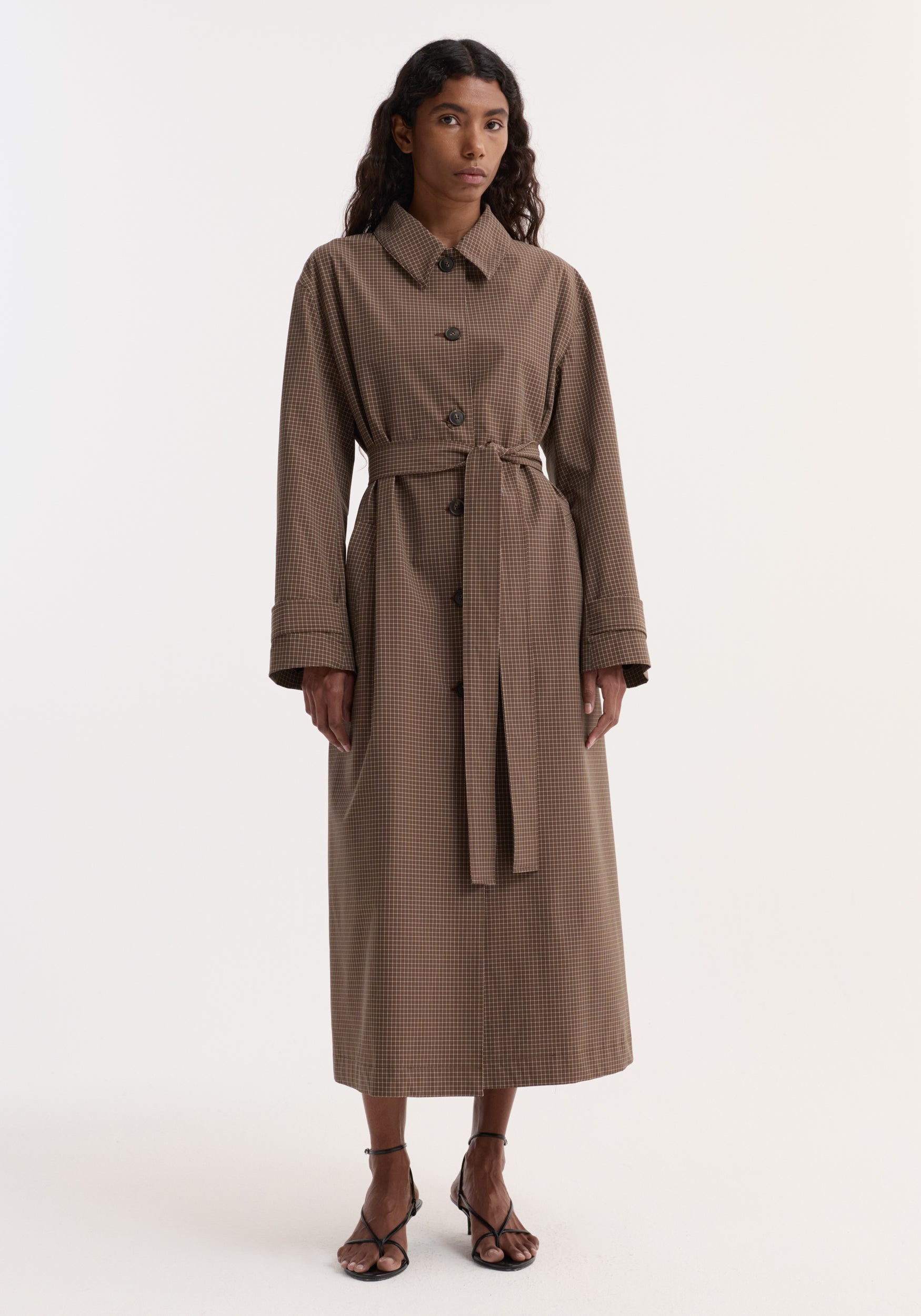 Woman wearing a long Technical check belted coat | brown multi check with button closure, wide collar, cuffed sleeves, and a matching belt tied at the waist, paired with open-toe heels.