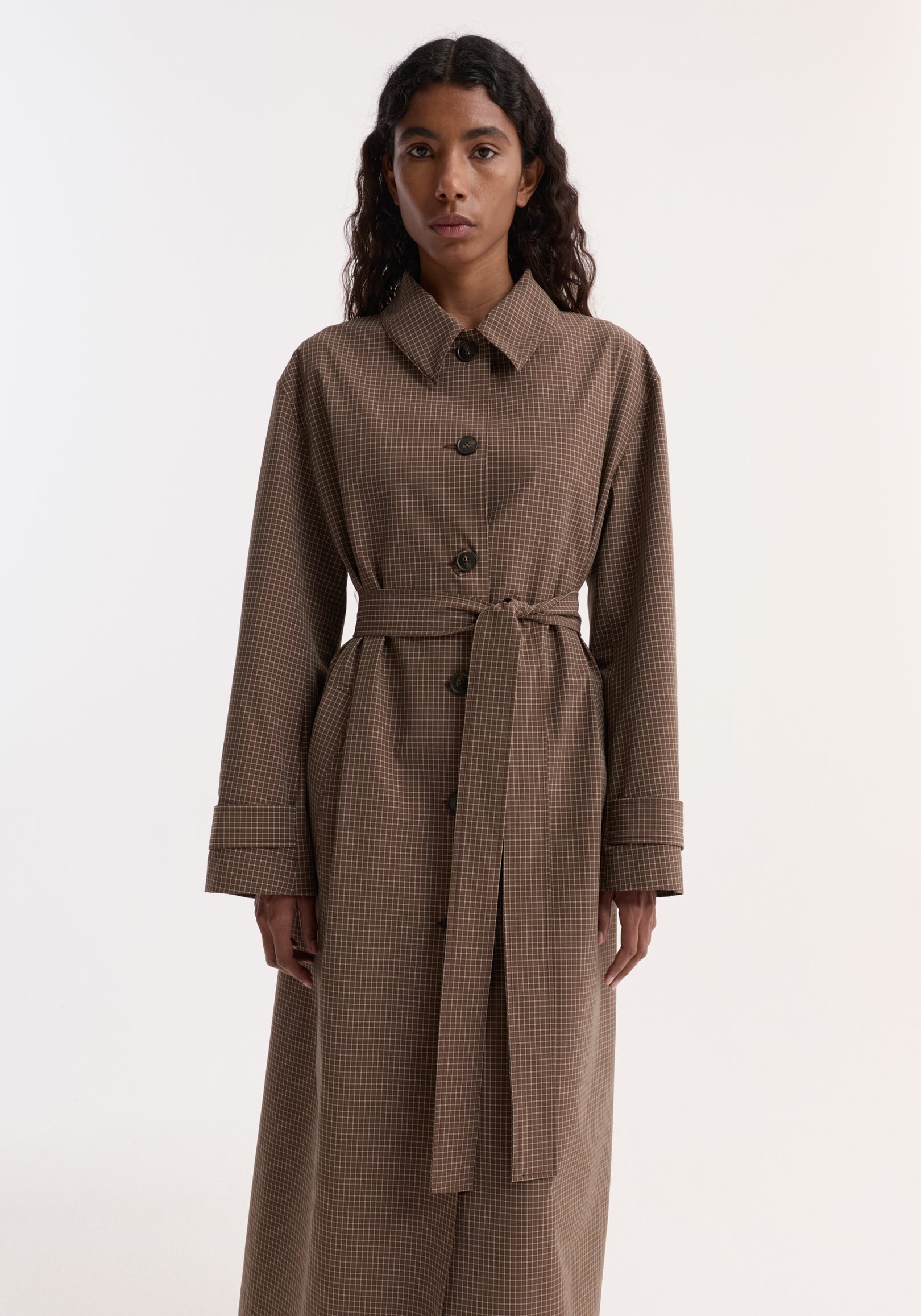 Woman wearing a long Technical check belted coat | brown multi check with a waist tie, buttons down the front, and long sleeves against a plain background.