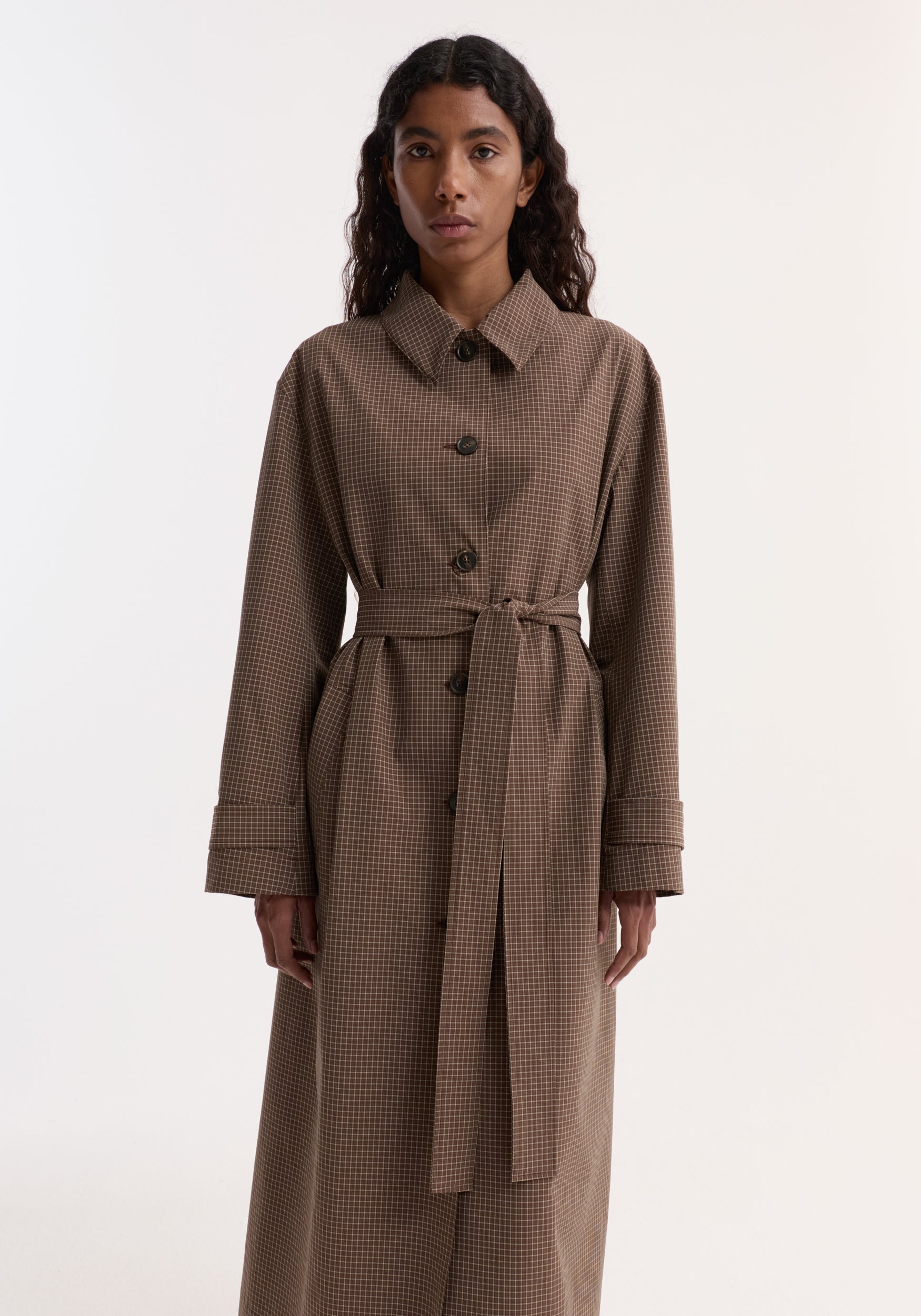 Woman wearing a long Technical check belted coat | brown multi check with a waist tie, buttons down the front, and long sleeves against a plain background.