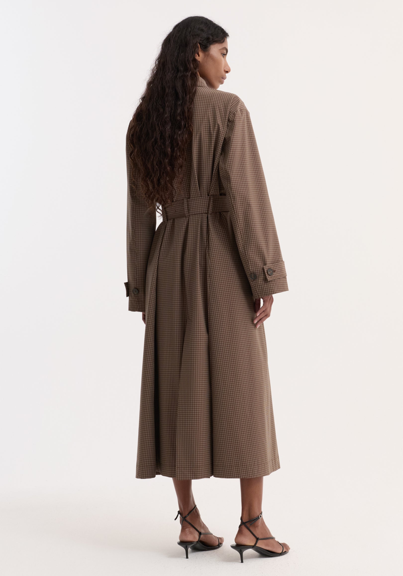 Woman wearing a long Technical check belted coat | brown multi check with a small check pattern, seen from the back, paired with black strappy heels against a plain white background.