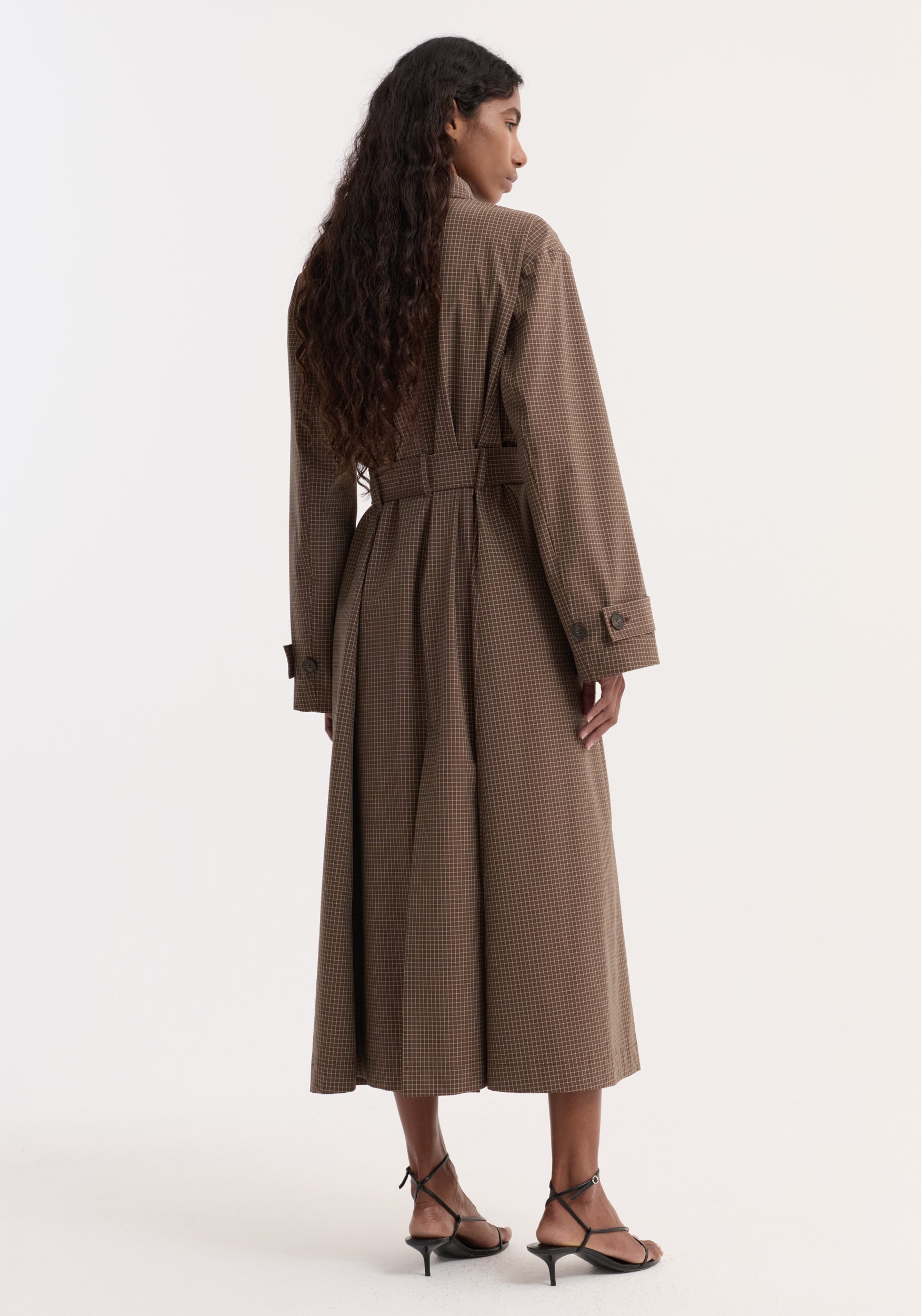 Woman wearing a long Technical check belted coat | brown multi check with a small check pattern, seen from the back, paired with black strappy heels against a plain white background.