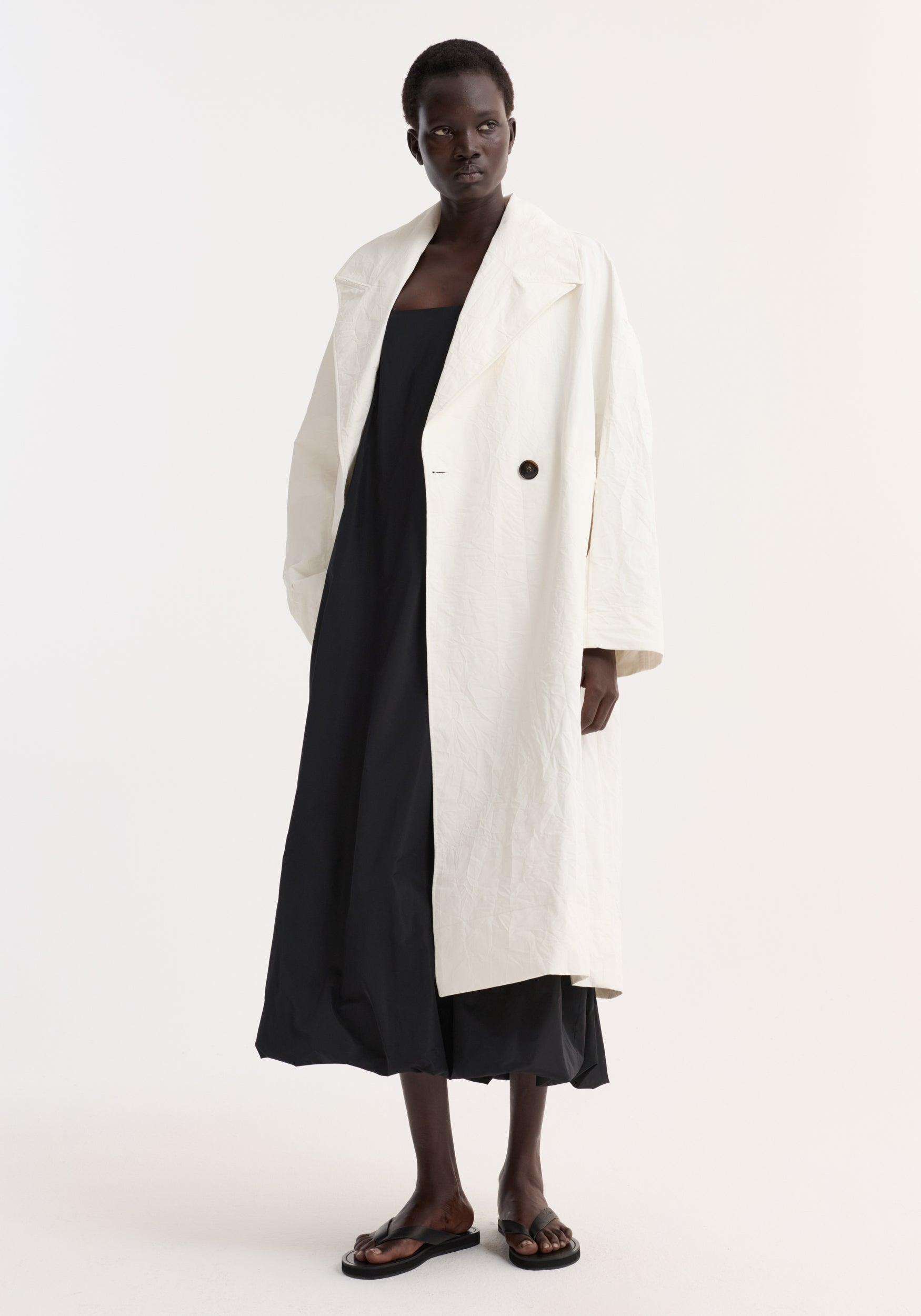A model wearing an oversized crushed cotton coat | off white over a long black dress and black sandals, standing against a plain light background.