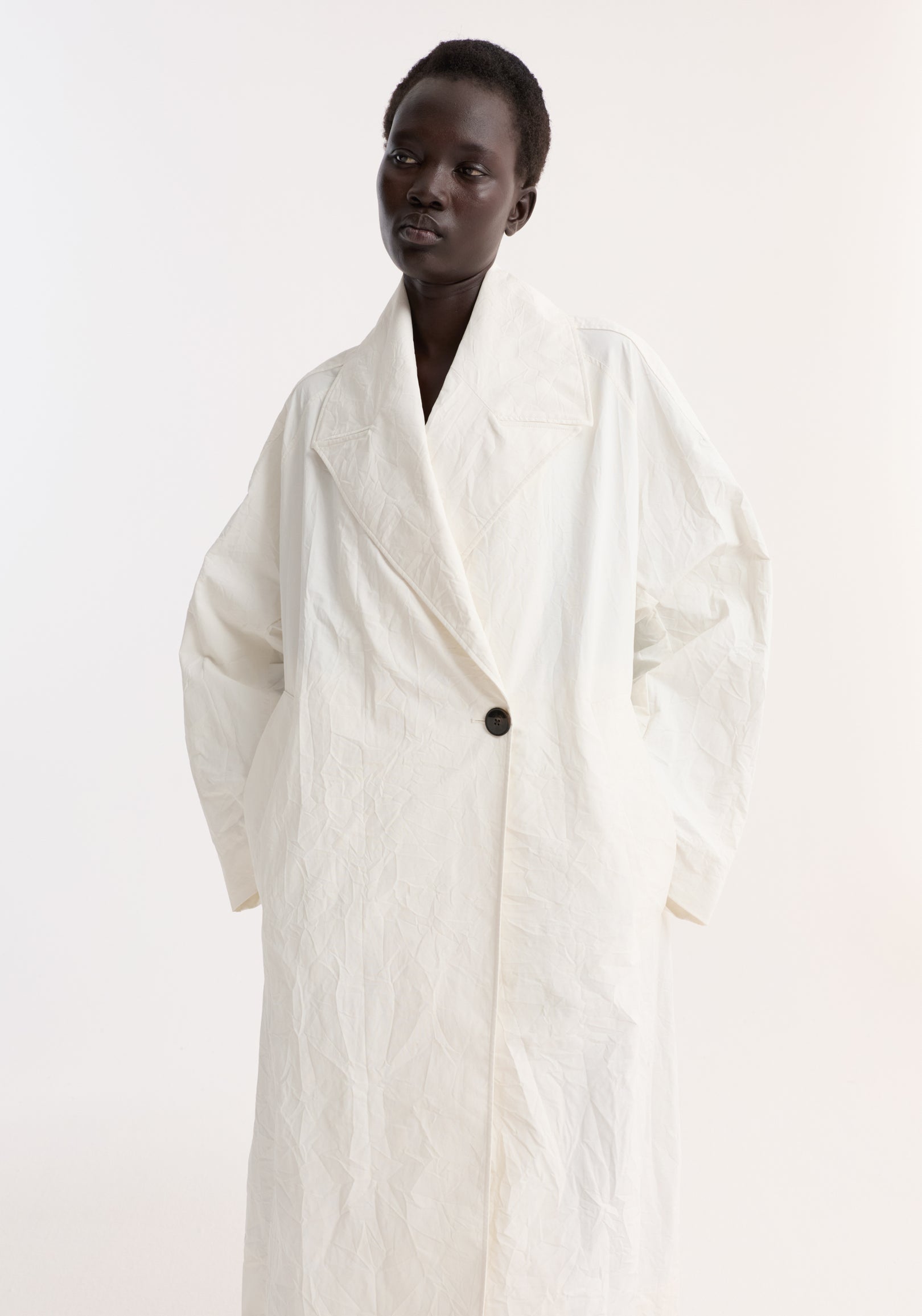 Model wearing an oversized crushed cotton coat in off white with a single black button and wide lapels against a plain light background, showcasing the textured fabric.