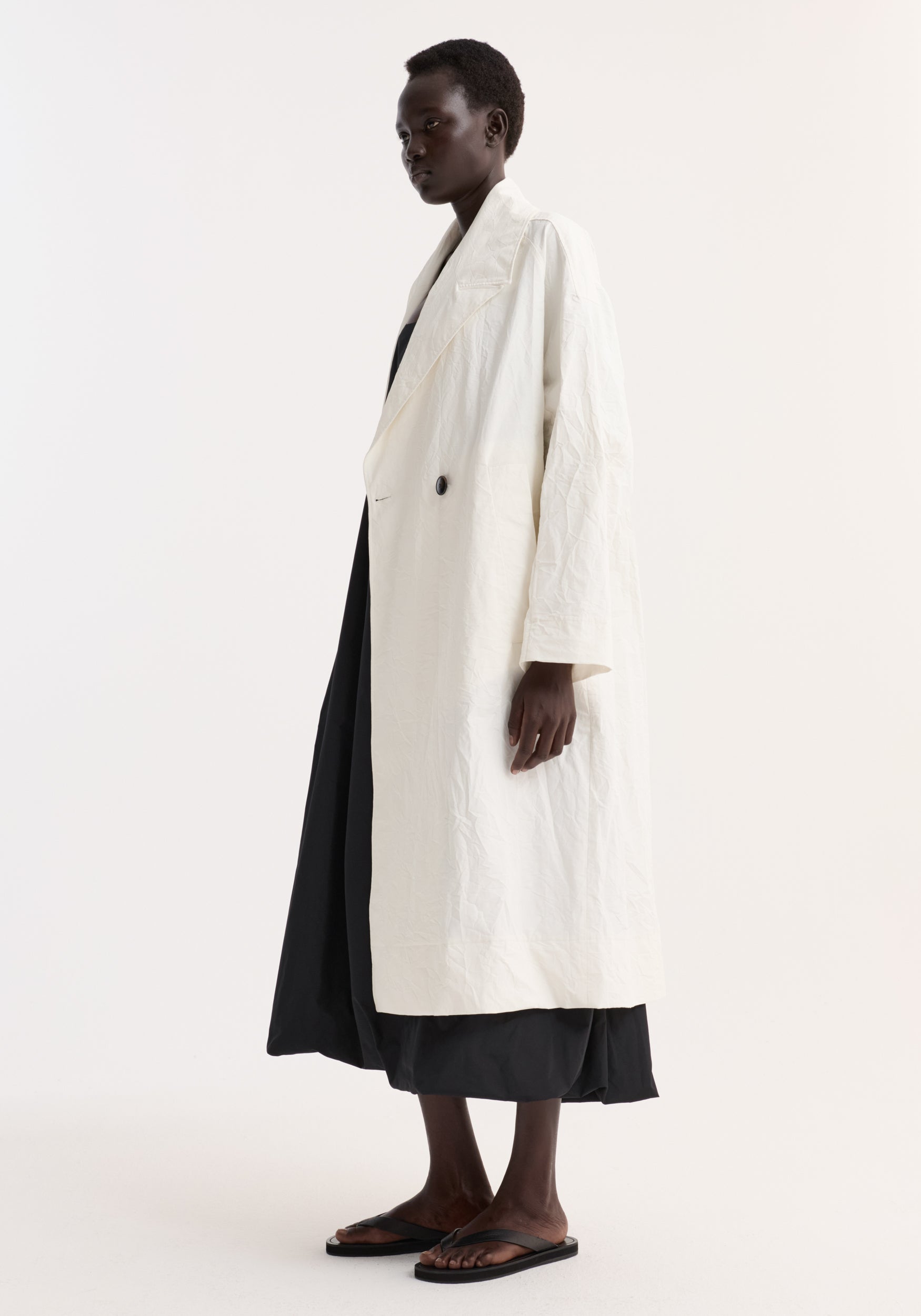 Side view of a model wearing an oversized crushed cotton coat | off white over a black dress and black sandals against a plain white background.