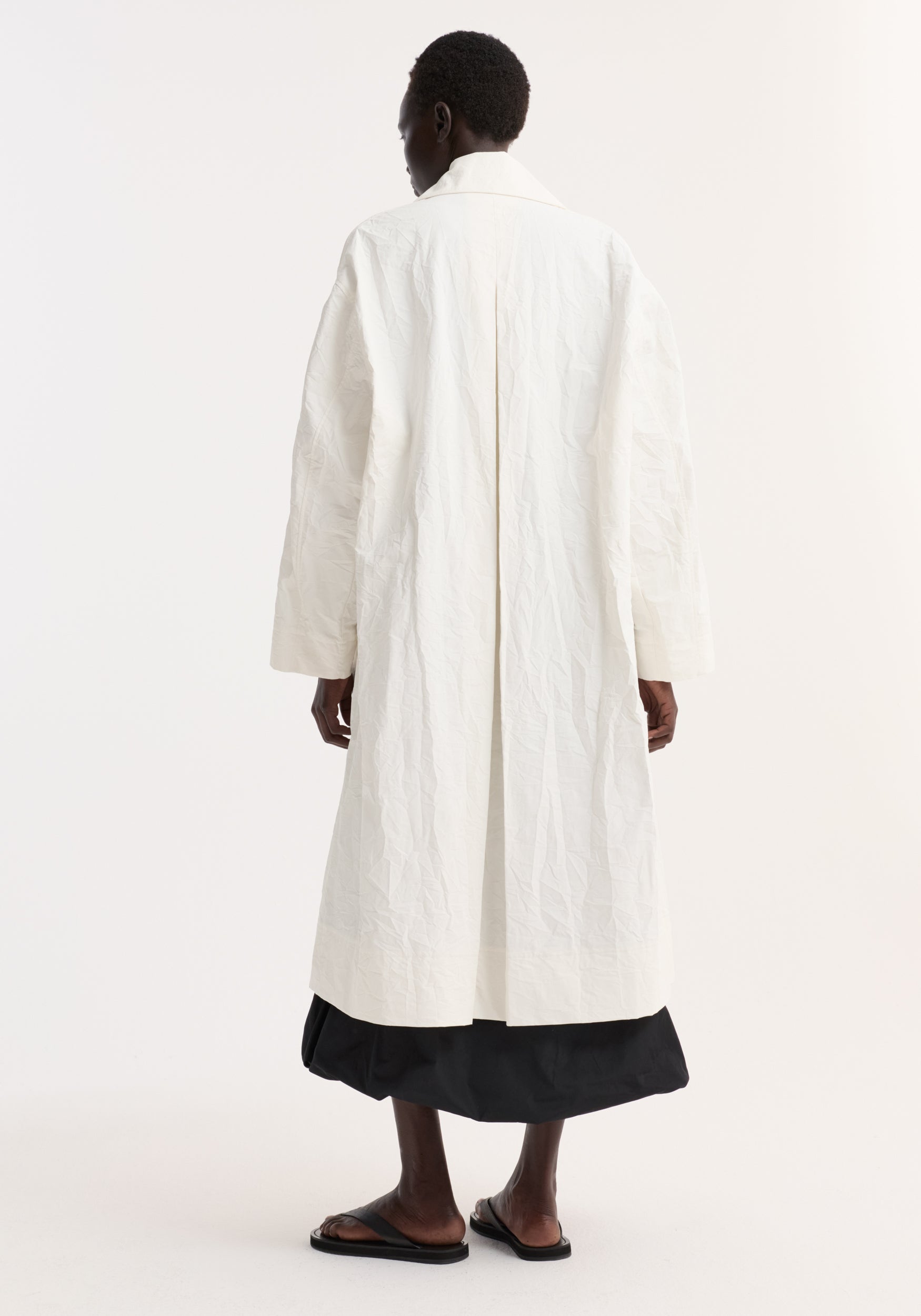 Back view of a model wearing an oversized crushed cotton coat | off white, featuring a relaxed fit and textured fabric, styled with black cropped pants and flat sandals.