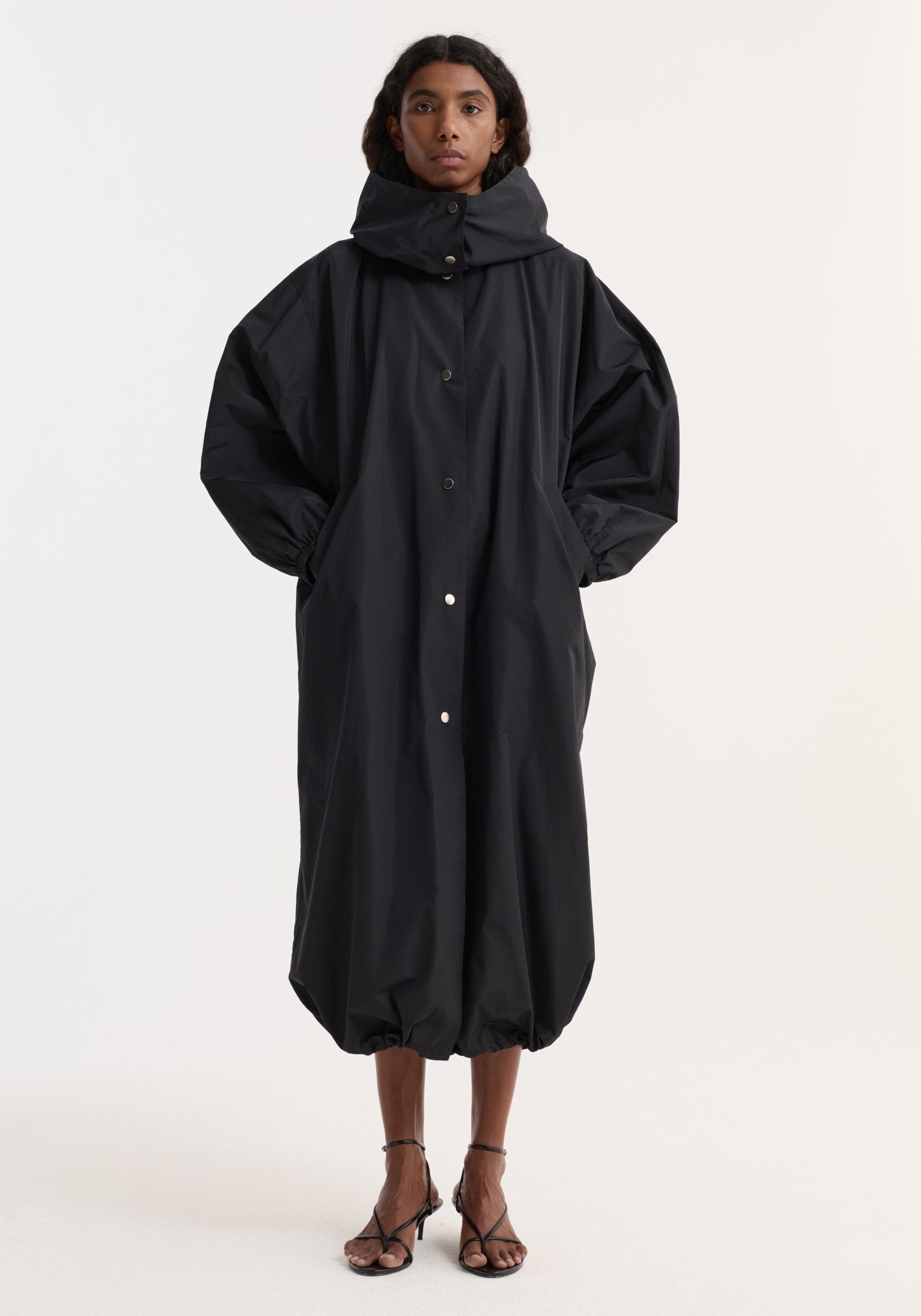 A model wearing a long, oversized black parachute silk-blend coat | black with a high collar, button-front closure, voluminous sleeves, and elasticized hem, paired with black strappy heels.