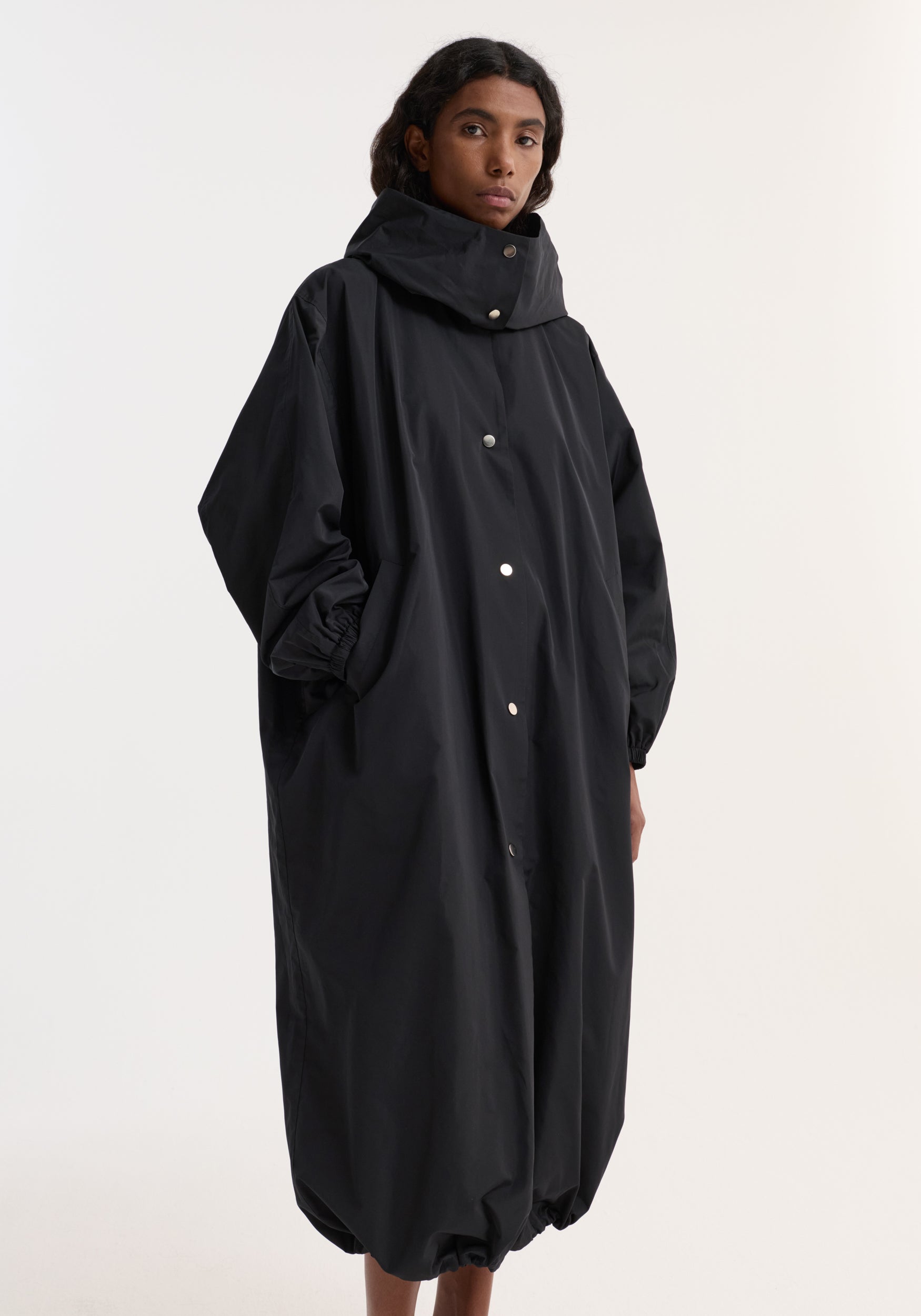 A model wearing a loose-fitting black parachute silk-blend coat with a high collar, snap button front, side pockets, and elasticized cuffs and hem, standing against a plain background.