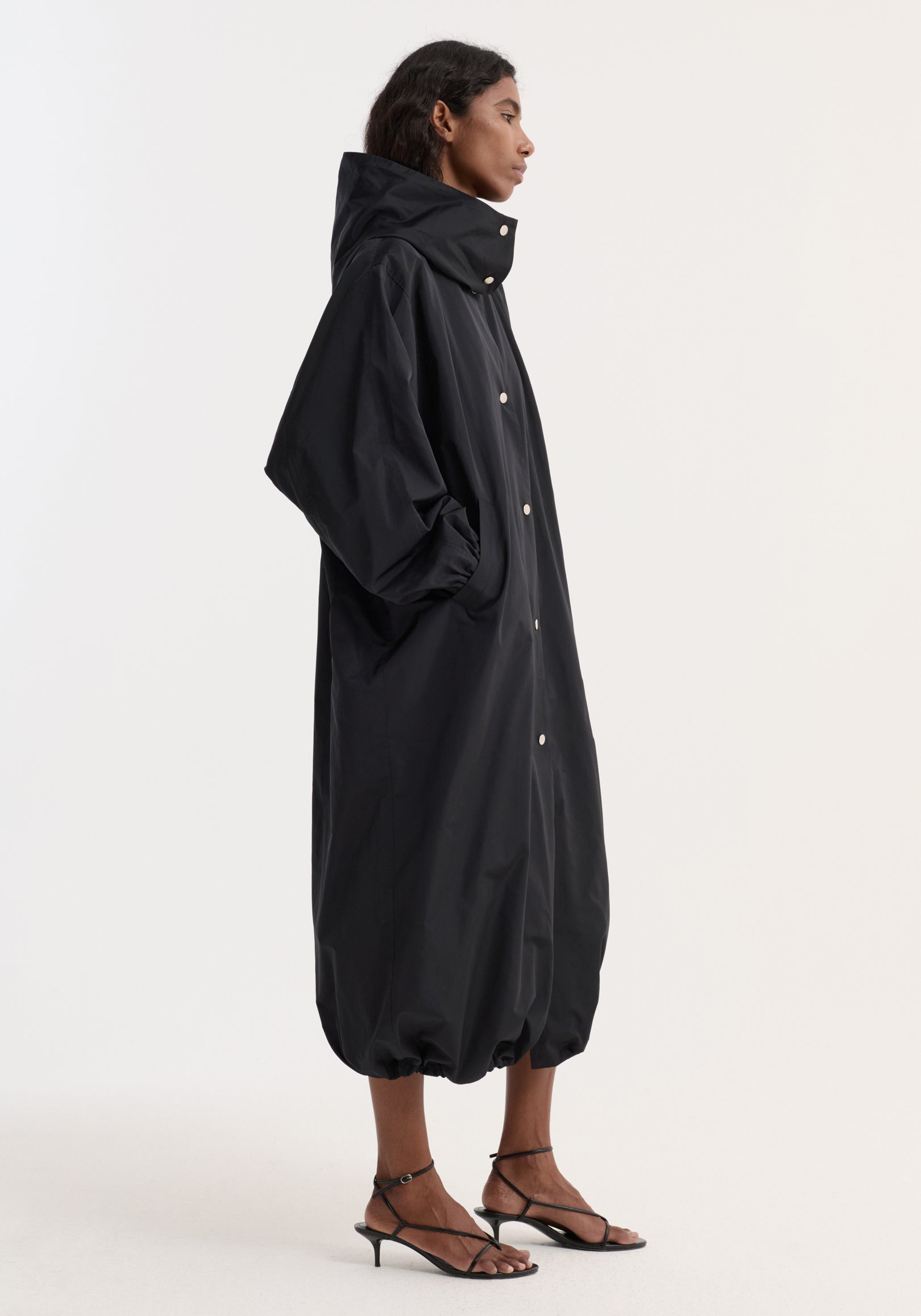 Side view of a woman wearing a long black Parachute silk-blend coat | black with a high collar, snap button closure, voluminous silhouette, and gathered hem, paired with black strappy heels.