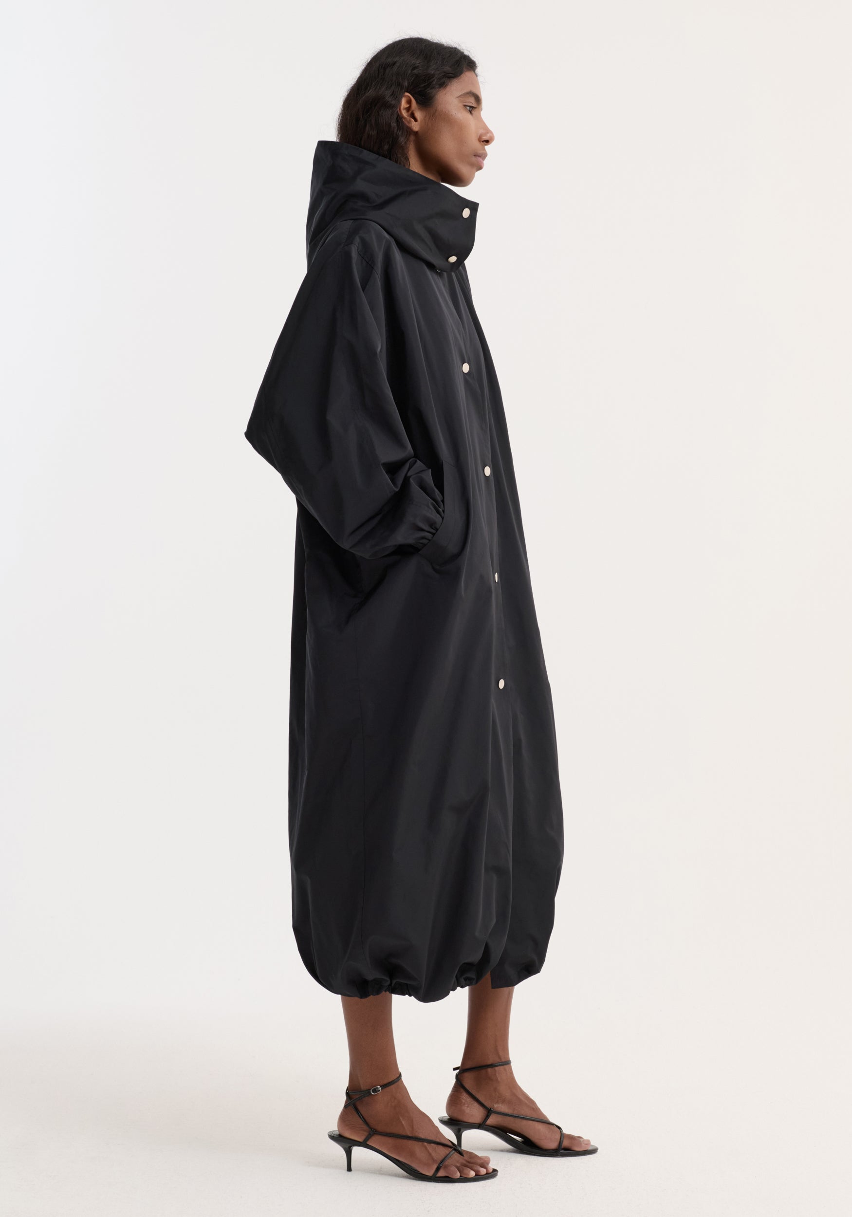 Side view of a woman wearing a long black Parachute silk-blend coat | black with a high collar, snap button closure, voluminous silhouette, and gathered hem, paired with black strappy heels.