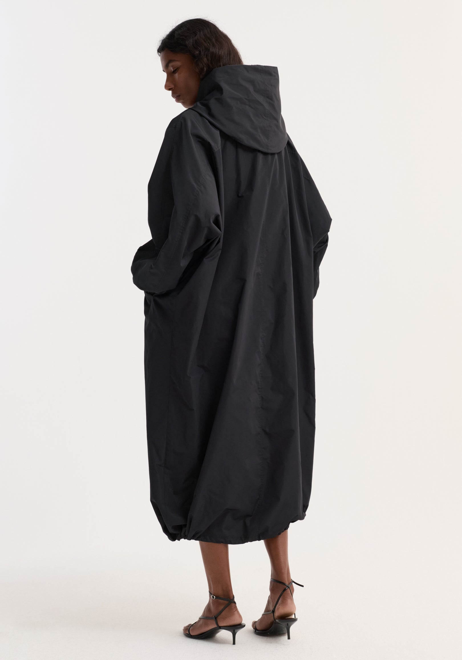 Woman wearing a long black parachute silk-blend coat with a hood, viewed from the back, paired with strappy black sandals.