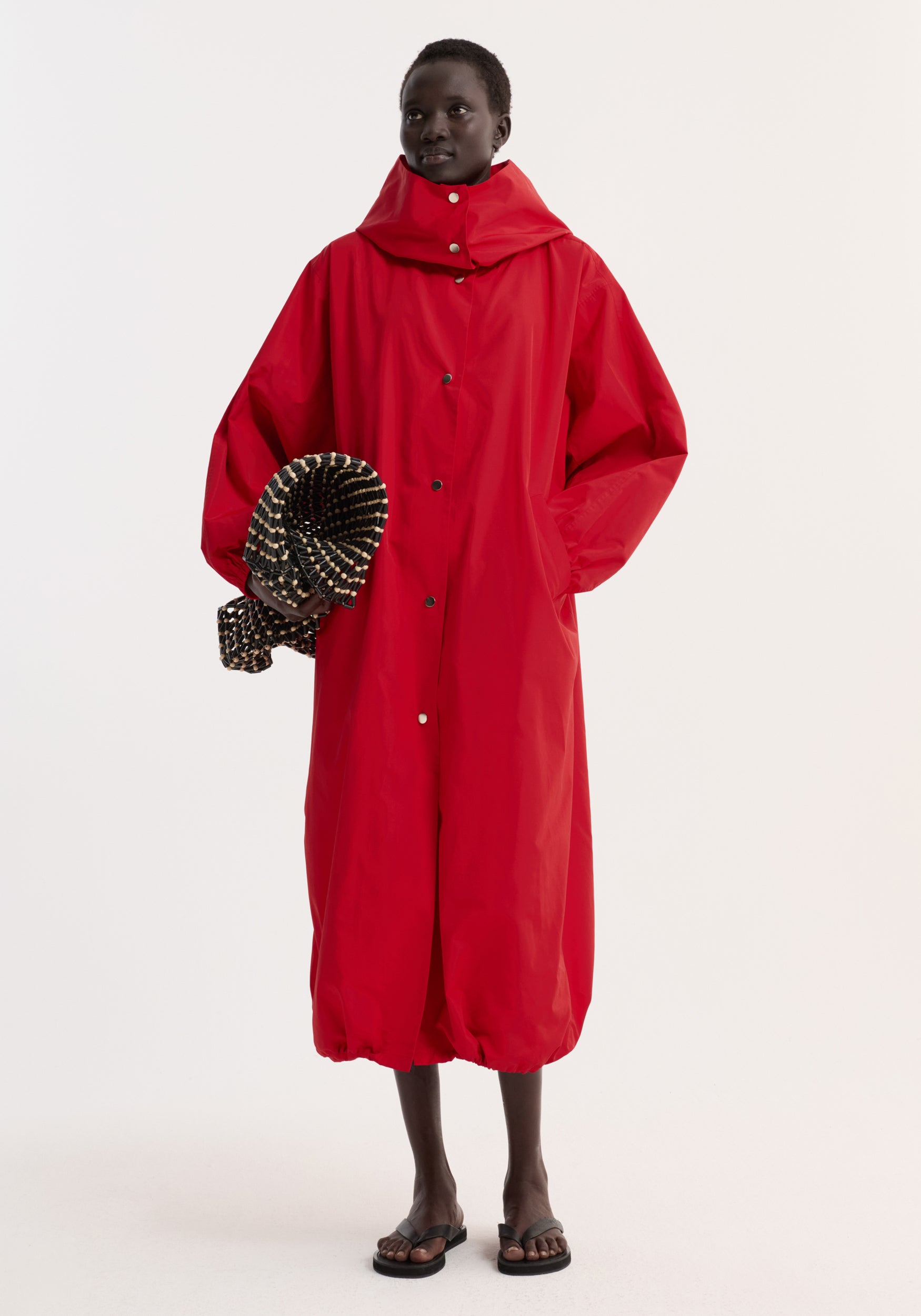 Model wearing a vibrant red parachute silk-blend coat | red with a high collar and snap button closure, paired with black sandals and holding a textured black and beige accessory.