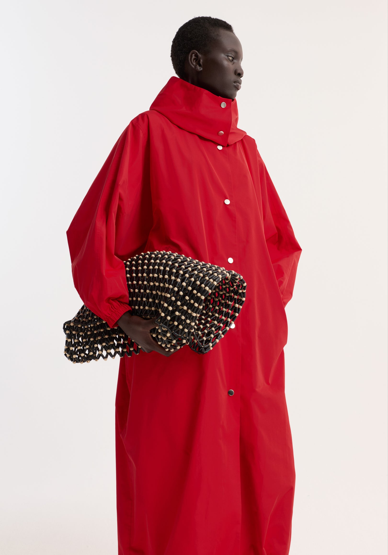 Model wearing a bright red Parachute silk-blend coat | red with a high collar and button closure, holding a black and beige woven clutch bag.