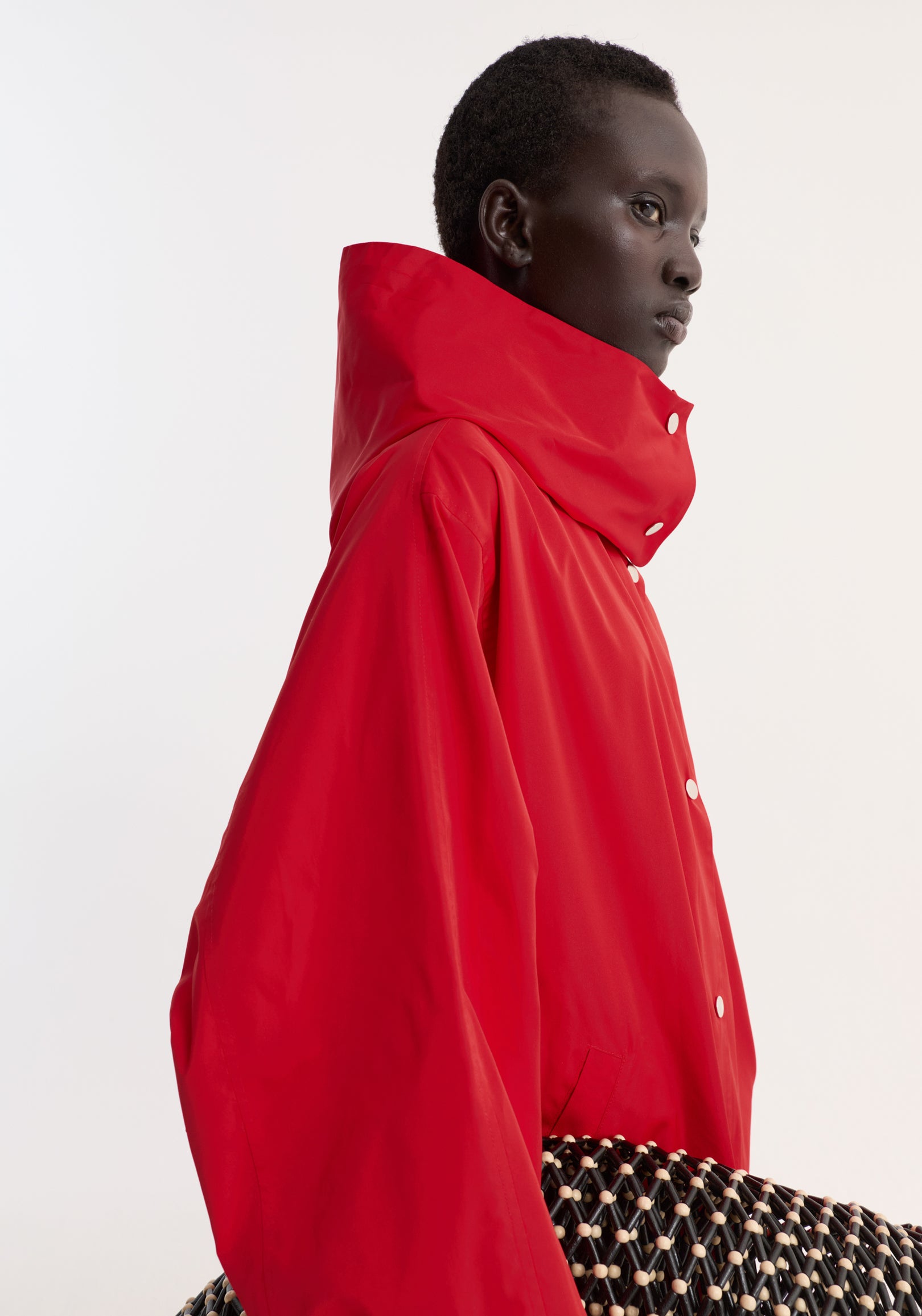 Profile view of a model wearing a vibrant parachute silk-blend coat | red with a high collar and snap buttons, holding a woven black and beige accessory.