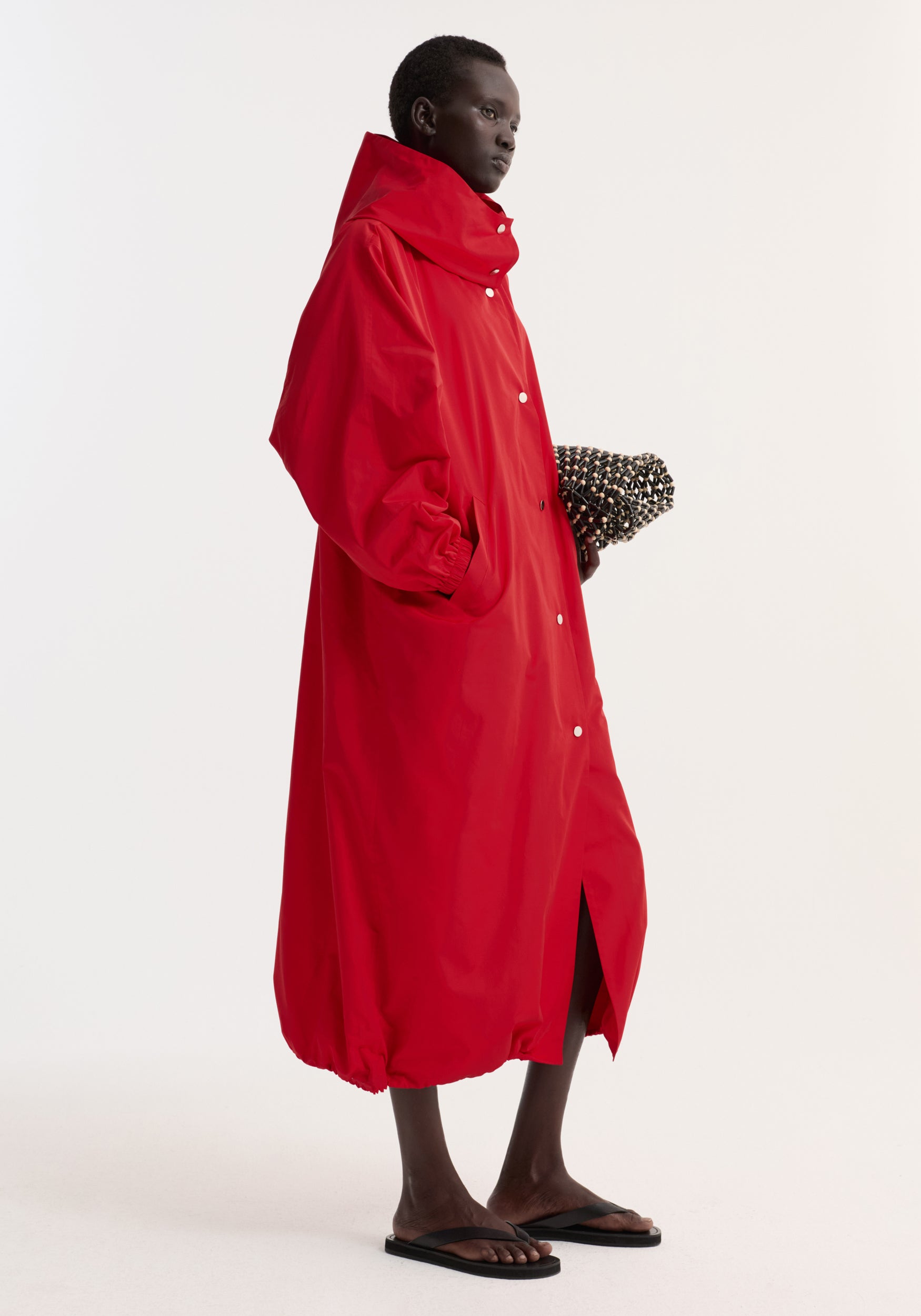 Model wearing a bright red parachute silk-blend coat | red with a high collar and button closures, styled with black sandals and holding a textured clutch bag.