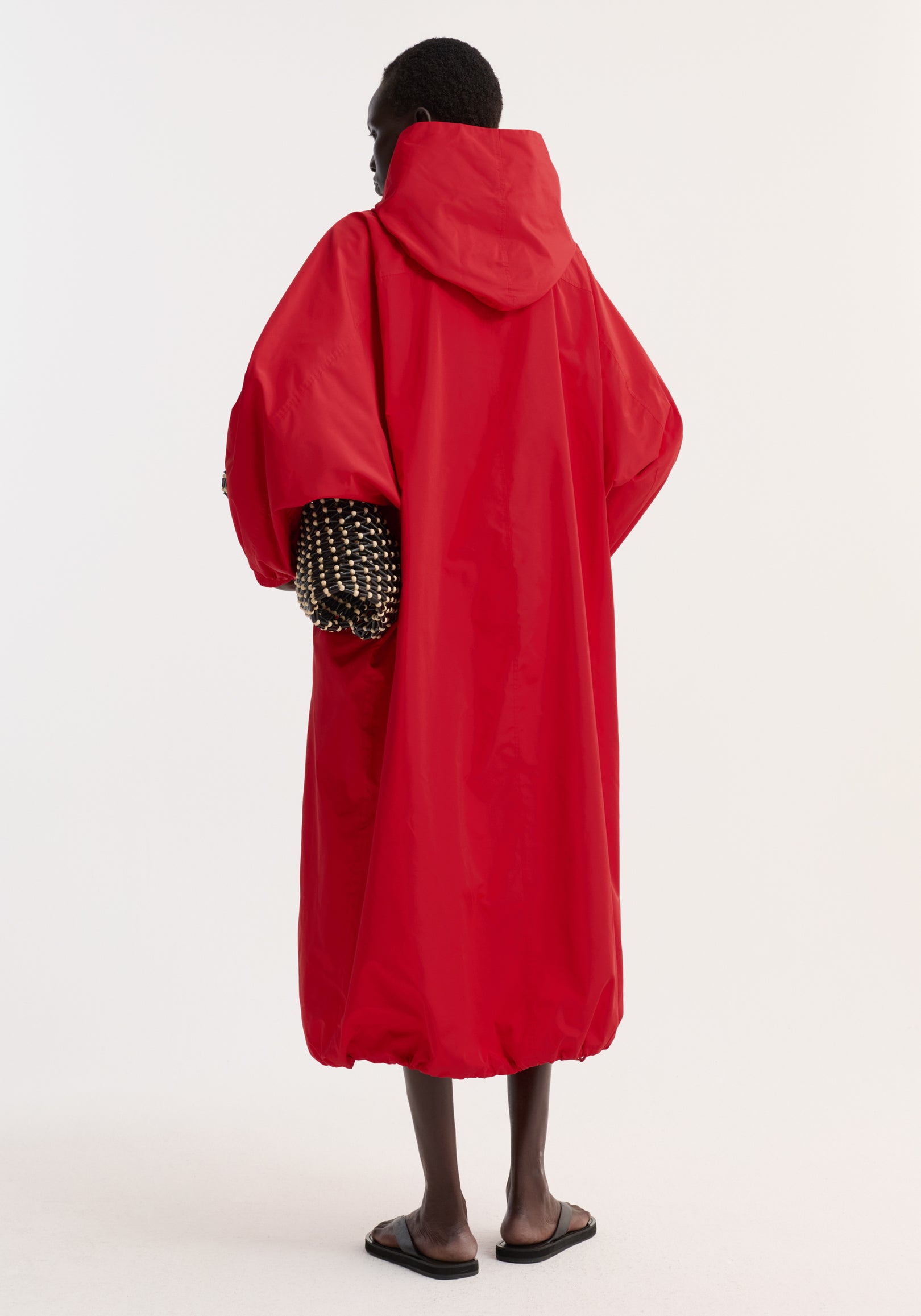 Person wearing a loose-fitting parachute silk-blend coat | red with a hood, viewed from the back, paired with black sandals and a patterned inner sleeve visible.