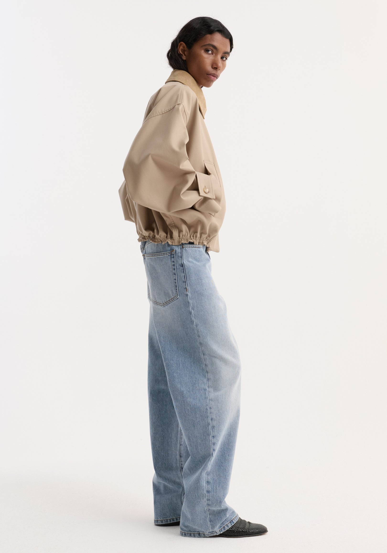 Woman wearing a suede collar blouson jacket | sand with elastic hem, paired with light blue wide-leg jeans and black shoes, standing in profile against a white background.