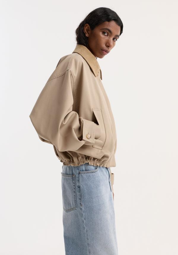 Side-profile of a model wearing a Suede collar blouson jacket | sand, showcasing its cropped, wide sleeves, and suede collar against a plain backdrop.