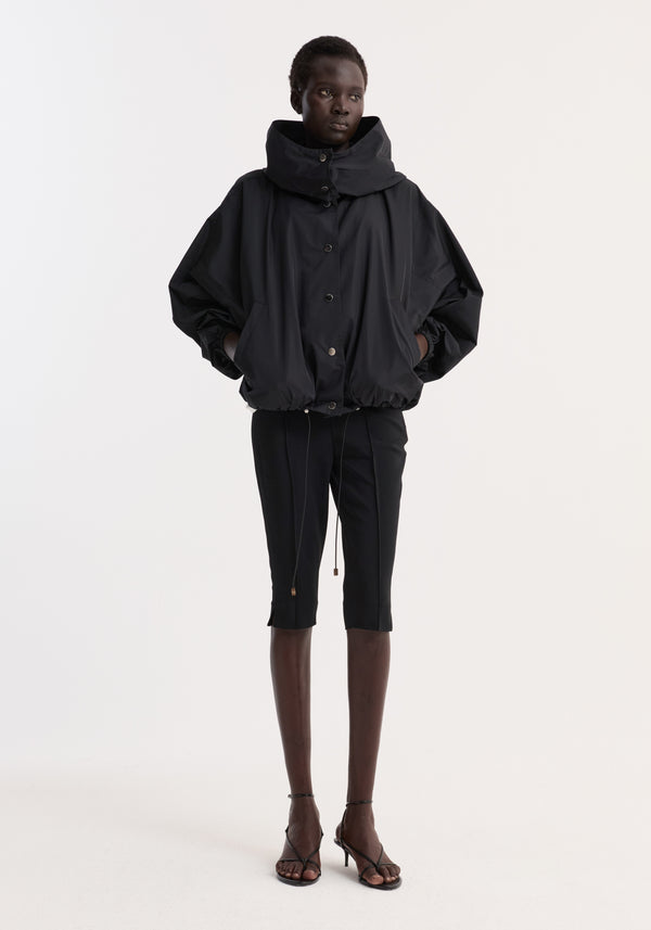 Dark-skinned model in Volume silk-blend jacket, featuring a high funnel collar, snap buttons, drawstring hem, over slim cropped pants and mid-heel sandals.