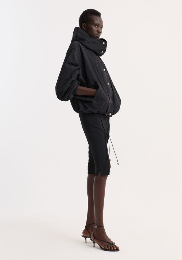 Volume silk-blend jacket in black, oversized with high collar, snap button closures, elasticated hem, worn by model in profile with hands in pockets.