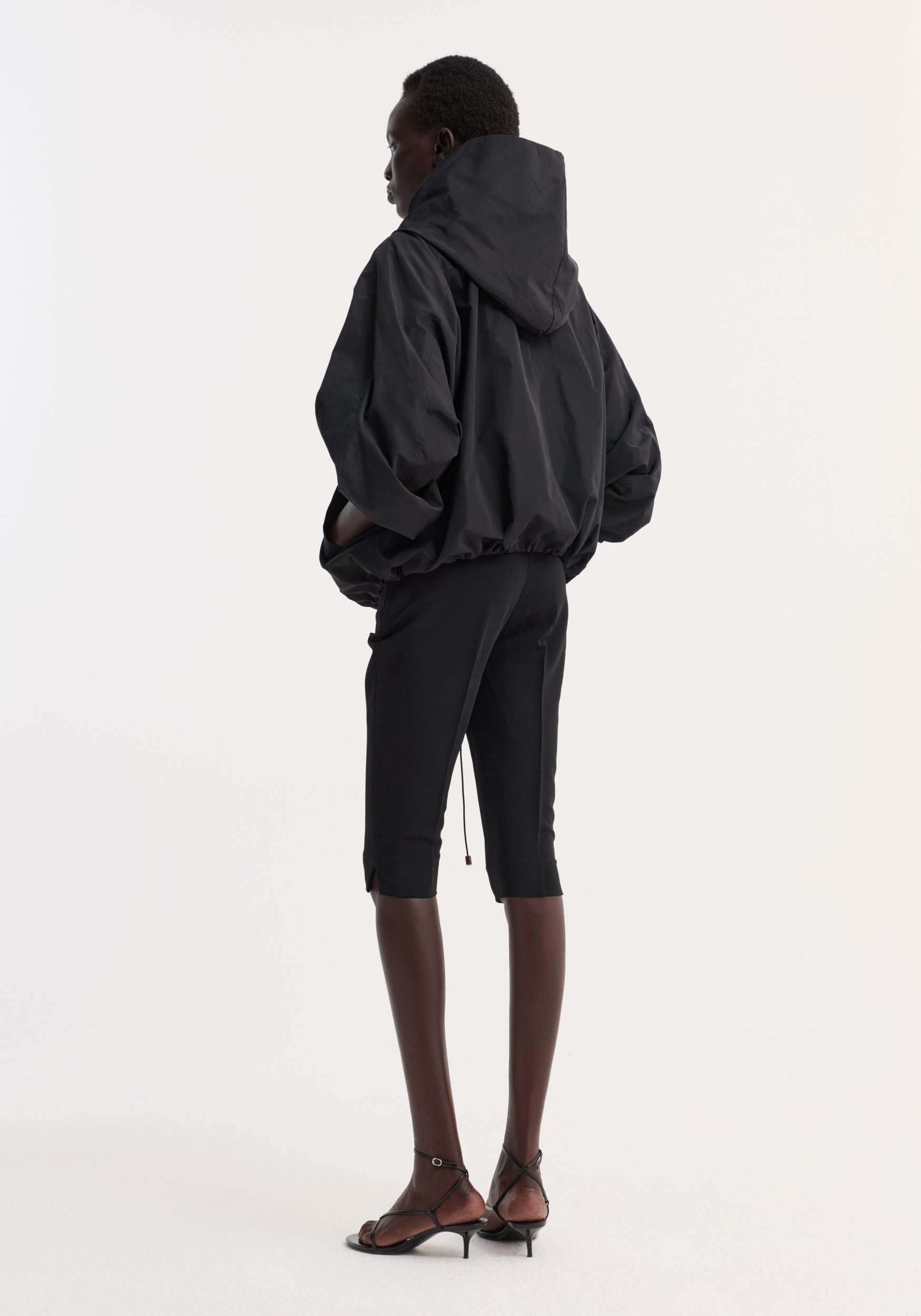 Model seen from behind wearing a black Volume silk-blend jacket | black with a hood, paired with fitted cropped pants and black strappy heels against a plain background.