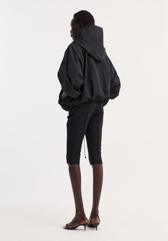 Model in Volume silk-blend jacket with generous hood, elasticated hem, and snap closures, paired with slim black pants and strappy sandals.