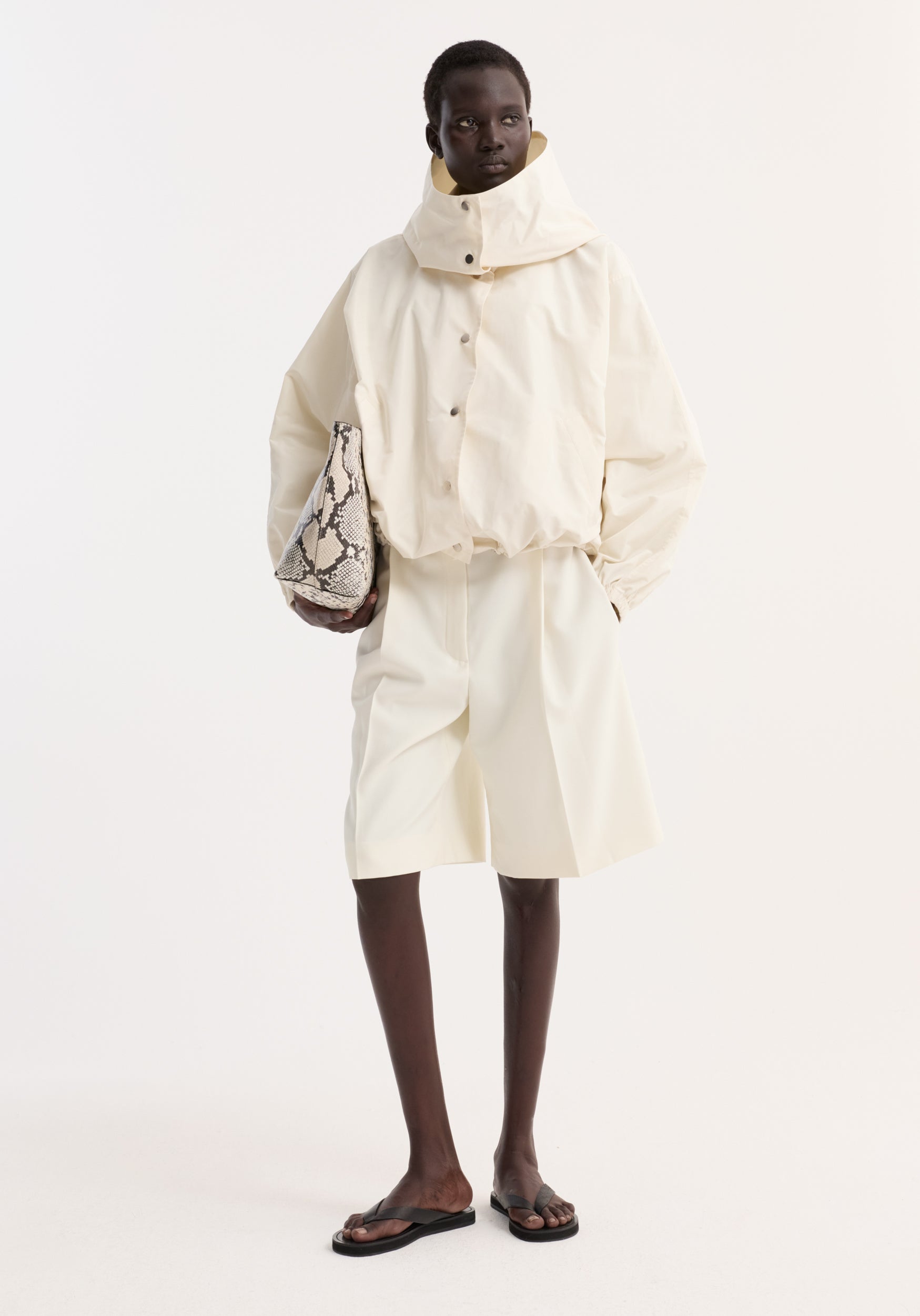 Model wearing a cream colored Volume silk-blend jacket | cream with a high collar and button closure, paired with matching wide-leg shorts and black sandals, holding a snakeskin-patterned bag.