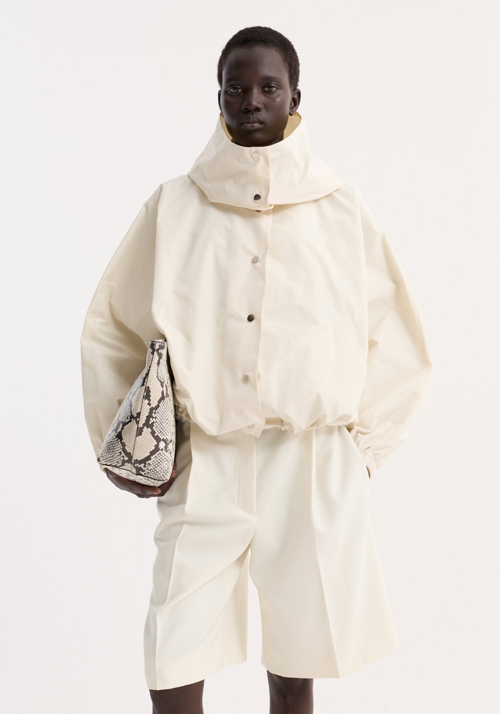 Model wearing a cream Volume silk-blend jacket with a high collar and snap buttons, paired with matching wide-leg shorts and holding a snakeskin-patterned bag.
