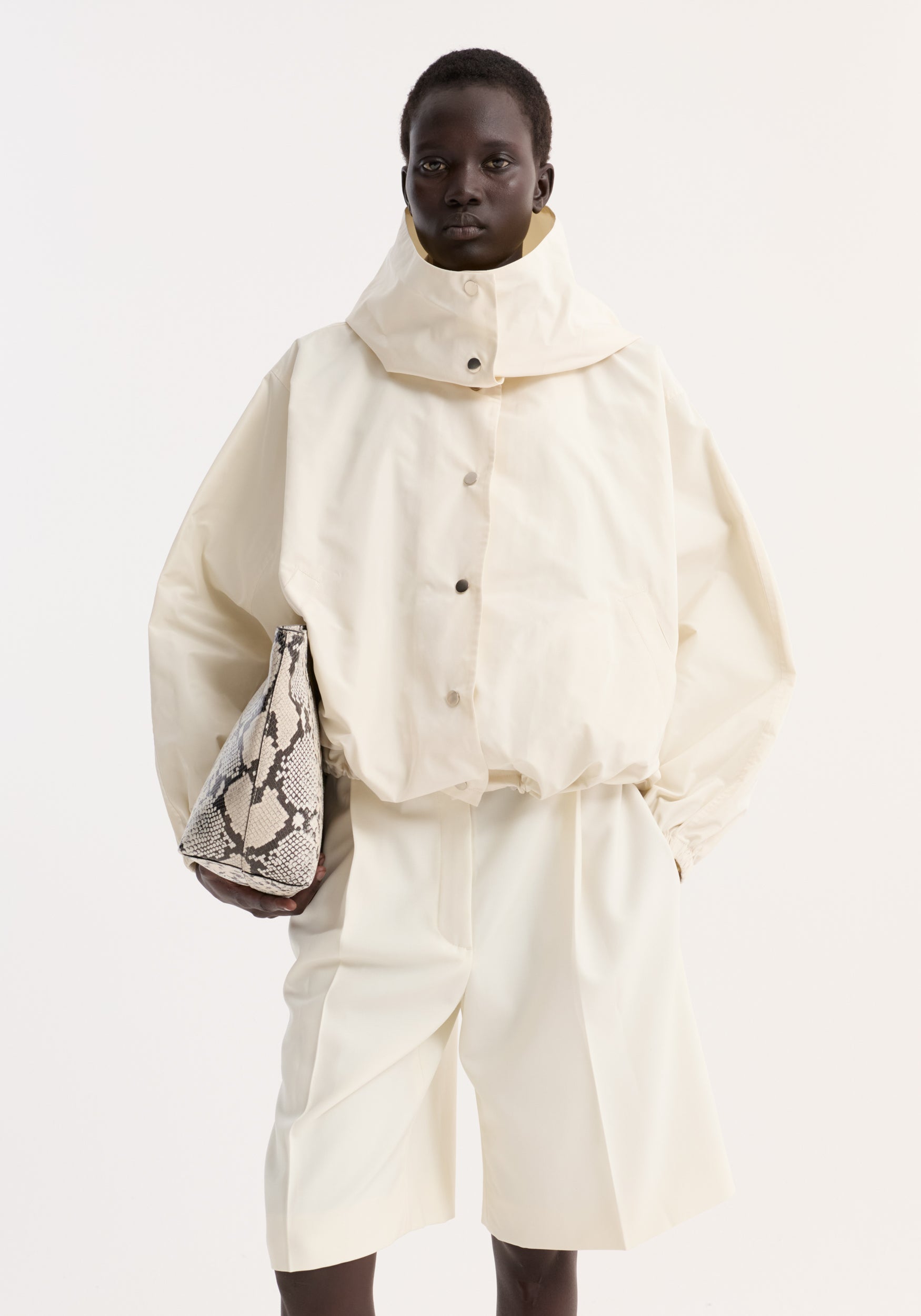 Model wearing a cream Volume silk-blend jacket with a high collar and snap buttons, paired with matching wide-leg shorts and holding a snakeskin-patterned bag.