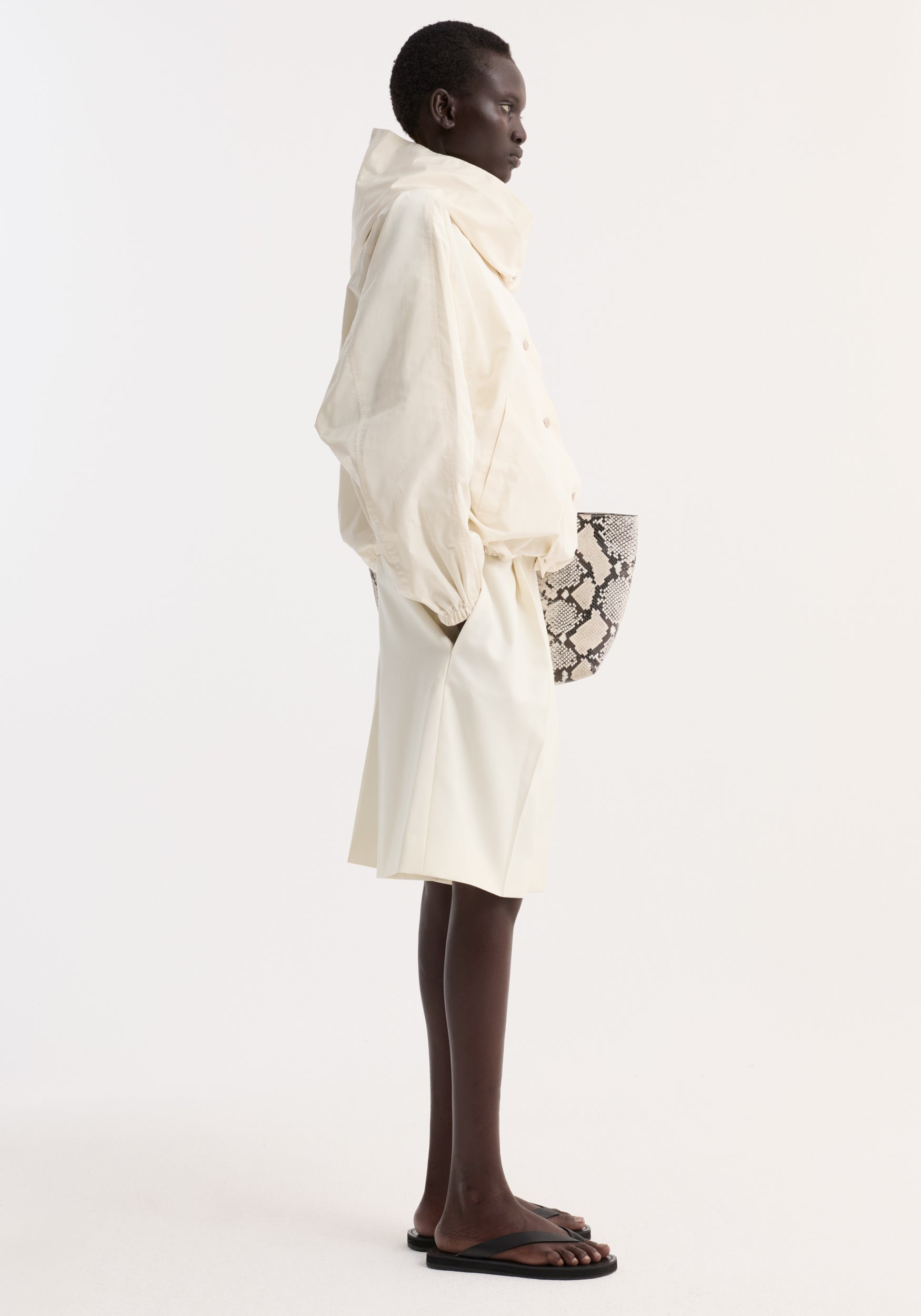 Side profile of a person wearing a cream Volume silk-blend jacket with a high collar and loose fit, paired with cream shorts and black sandals, holding a snakeskin-patterned bag.