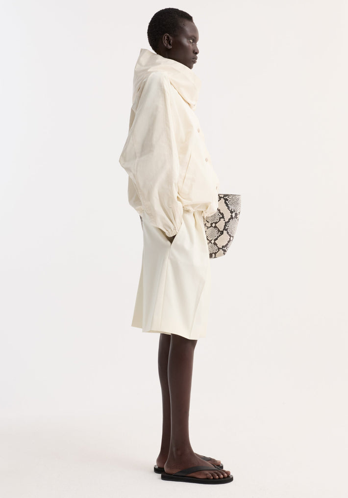 Model in profile wearing Volume silk-blend jacket | cream with hood, snap buttons, elasticated cuffs, over matching skirt, and carrying snakeskin print tote.