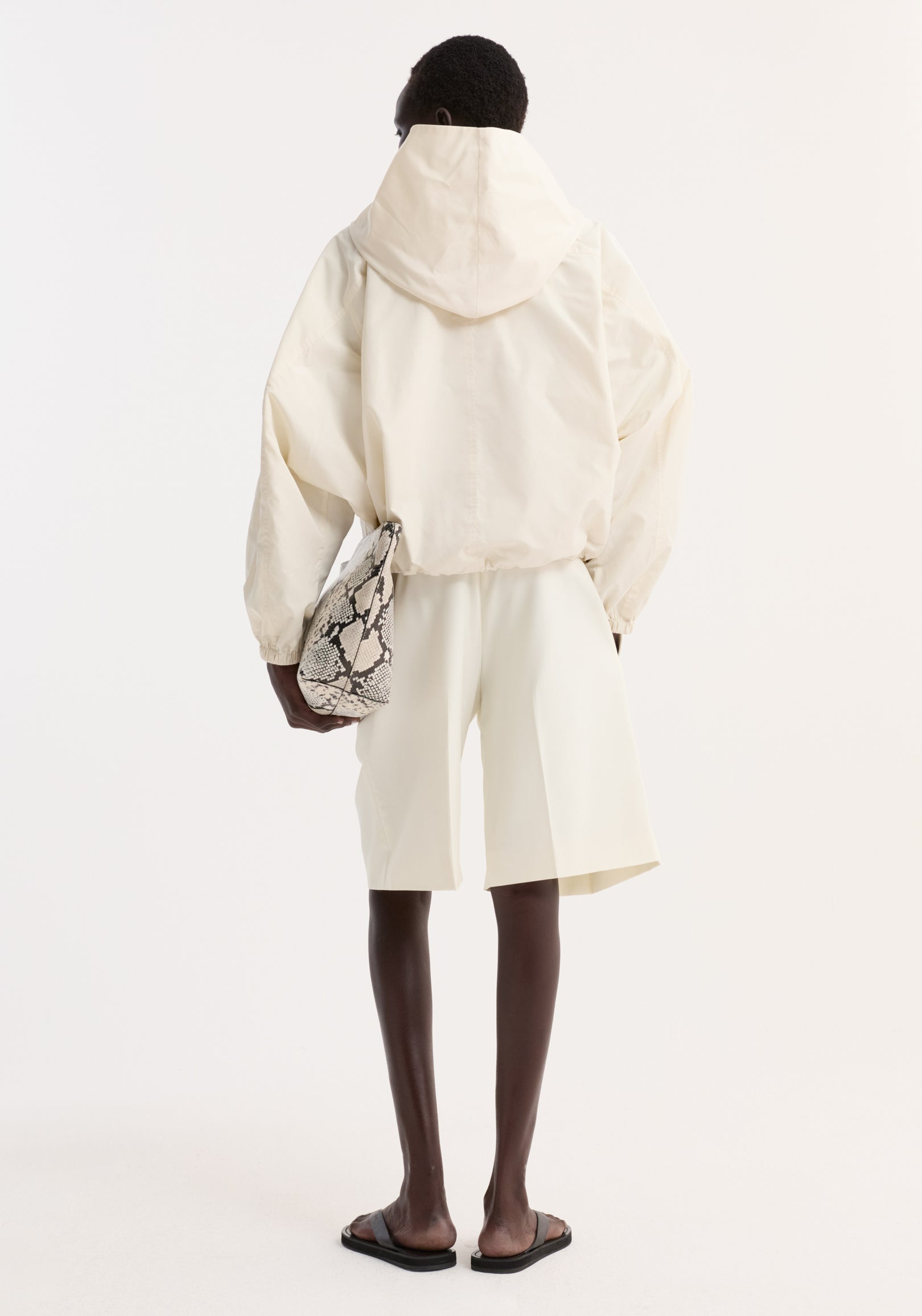 Back view of a person wearing a cream Volume silk-blend jacket with a hood, paired with matching cream shorts, black sandals, and holding a snake-patterned bag.