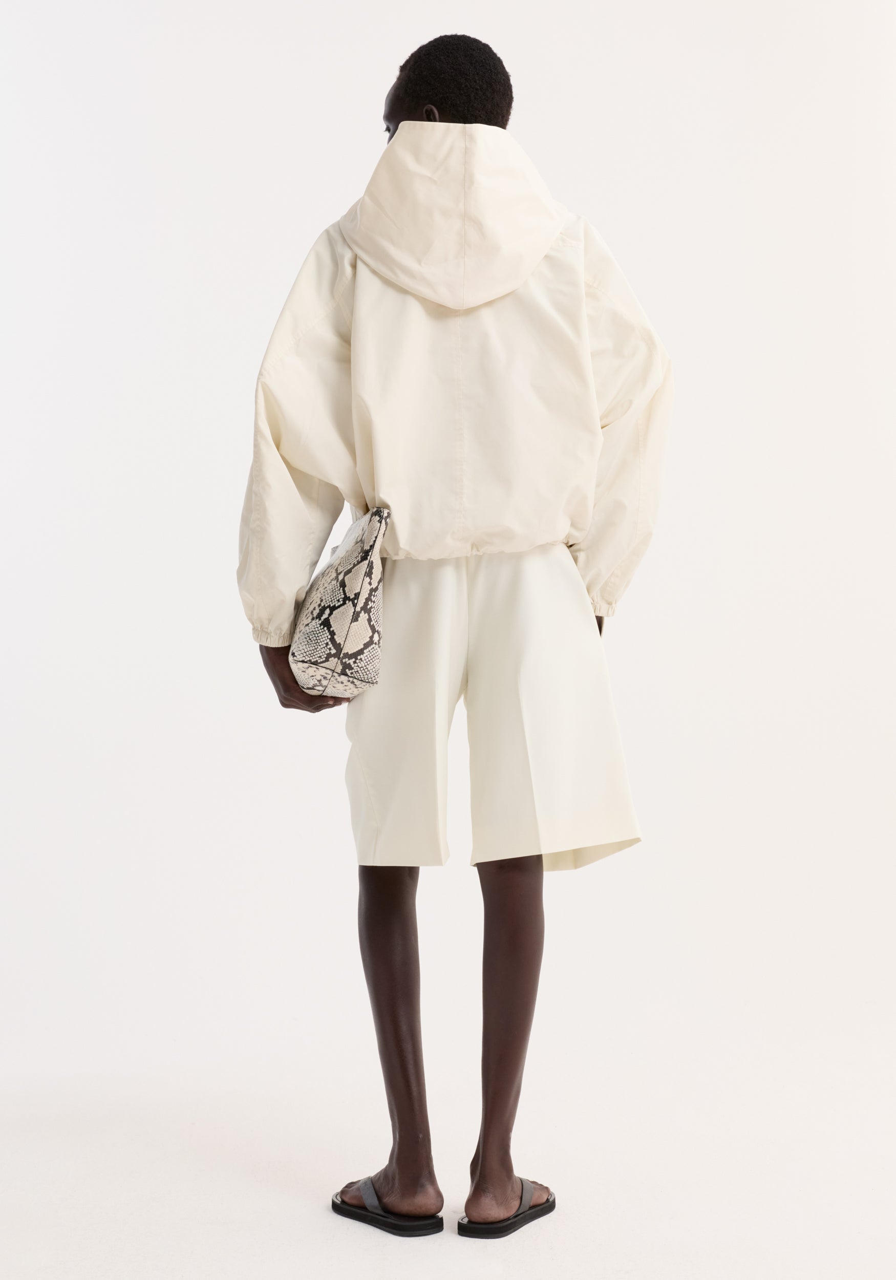 Back view of a person wearing a cream Volume silk-blend jacket with a hood, paired with matching cream shorts, black sandals, and holding a snake-patterned bag.
