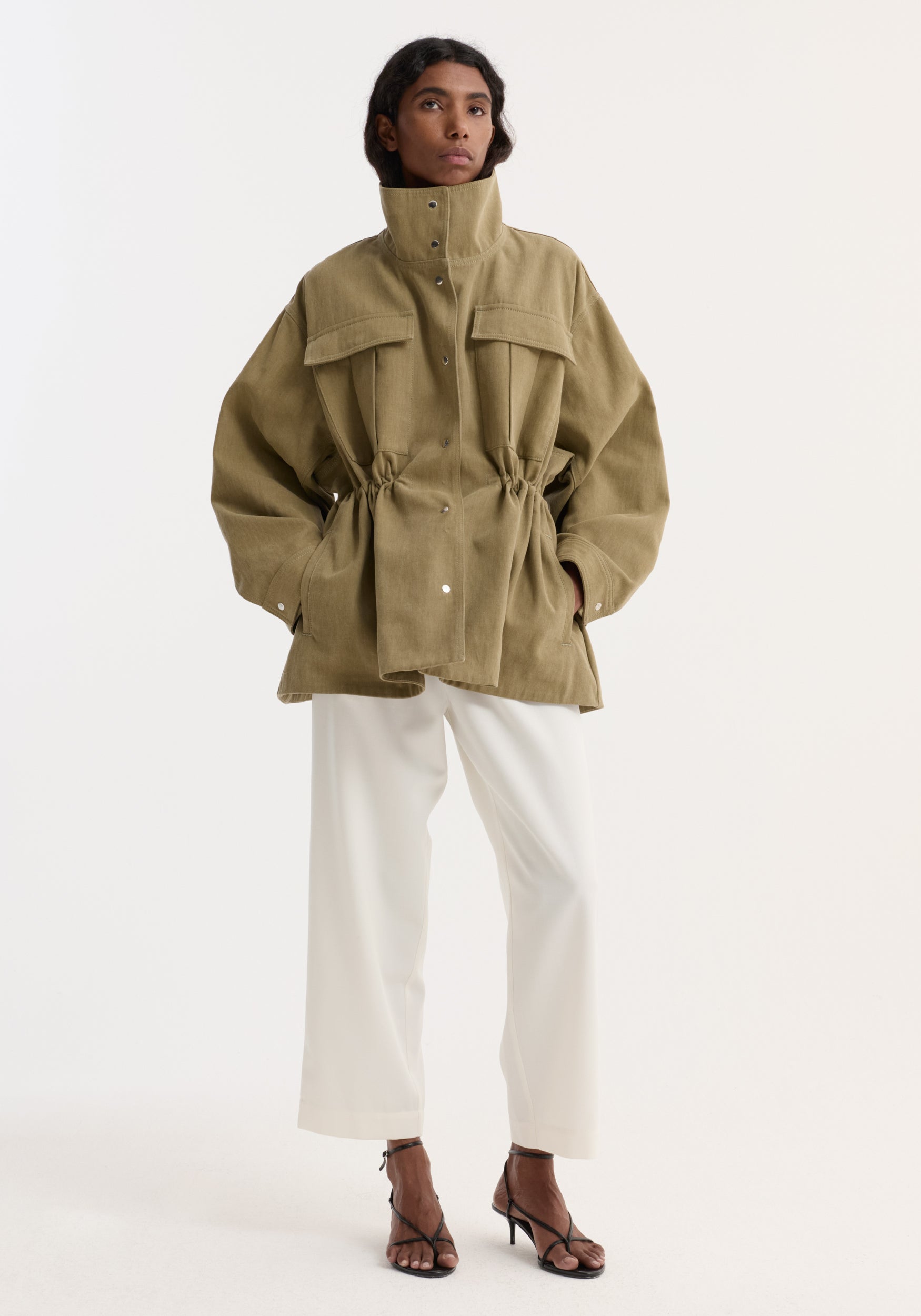 Model wearing a sculptural safari jacket | sage with high collar, flap pockets, drawstring waist, paired with white wide-leg pants and black heeled sandals against a plain background.