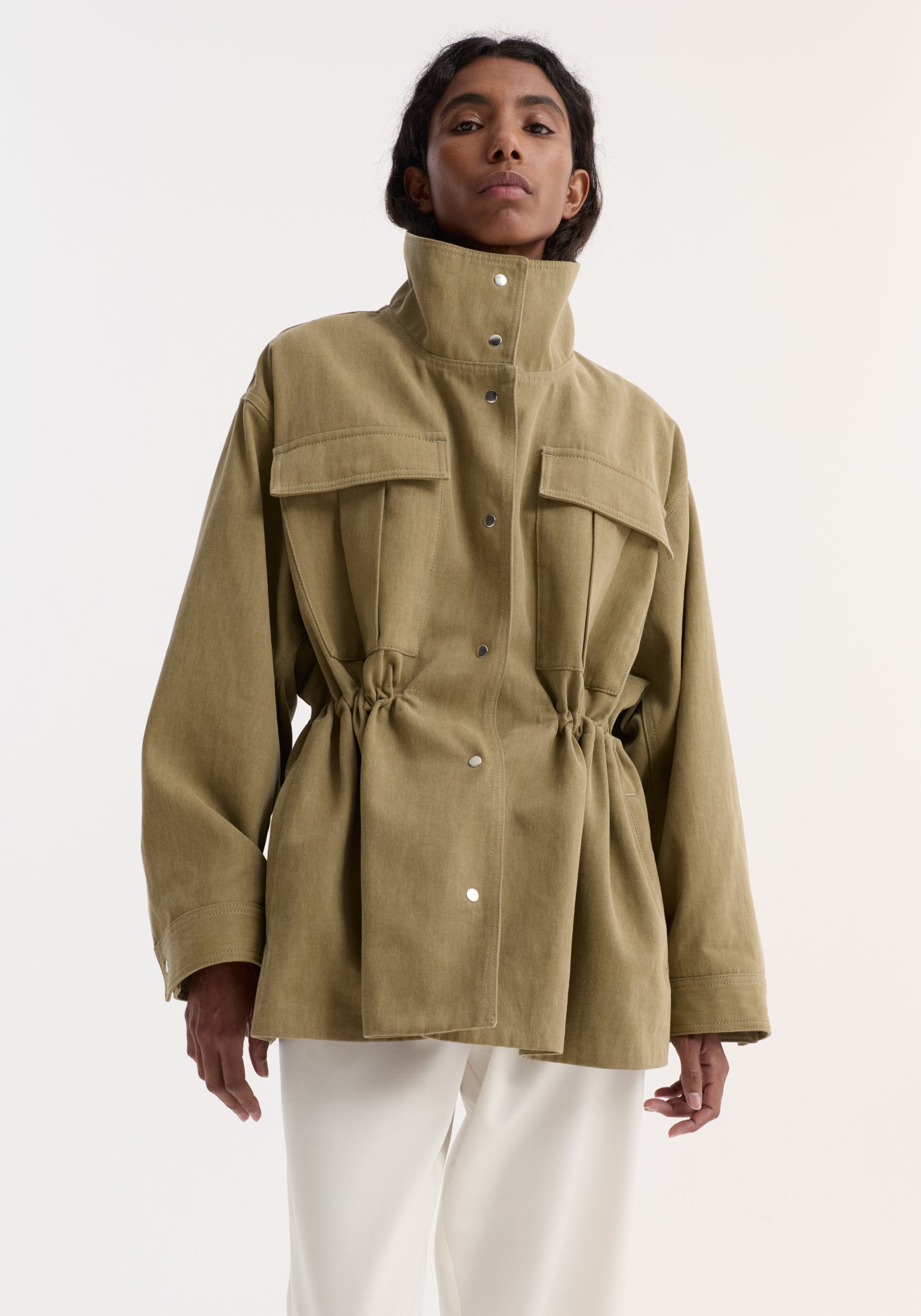 Woman wearing a sculptural safari jacket | sage with high collar, button closure, flap pockets, and cinched waist, paired with white pants against a plain background.