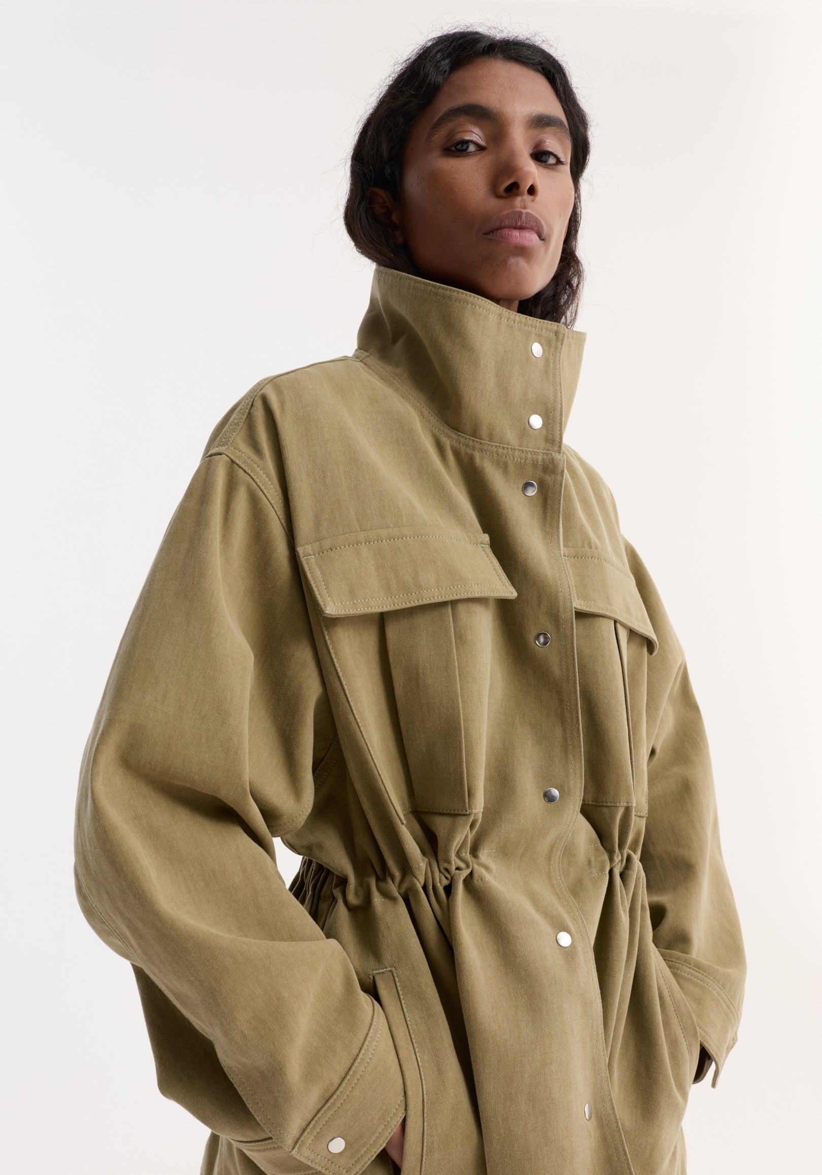Model wearing a sage colored sculptural safari jacket with high collar, snap buttons, front flap pockets, and cinched waist detailing.