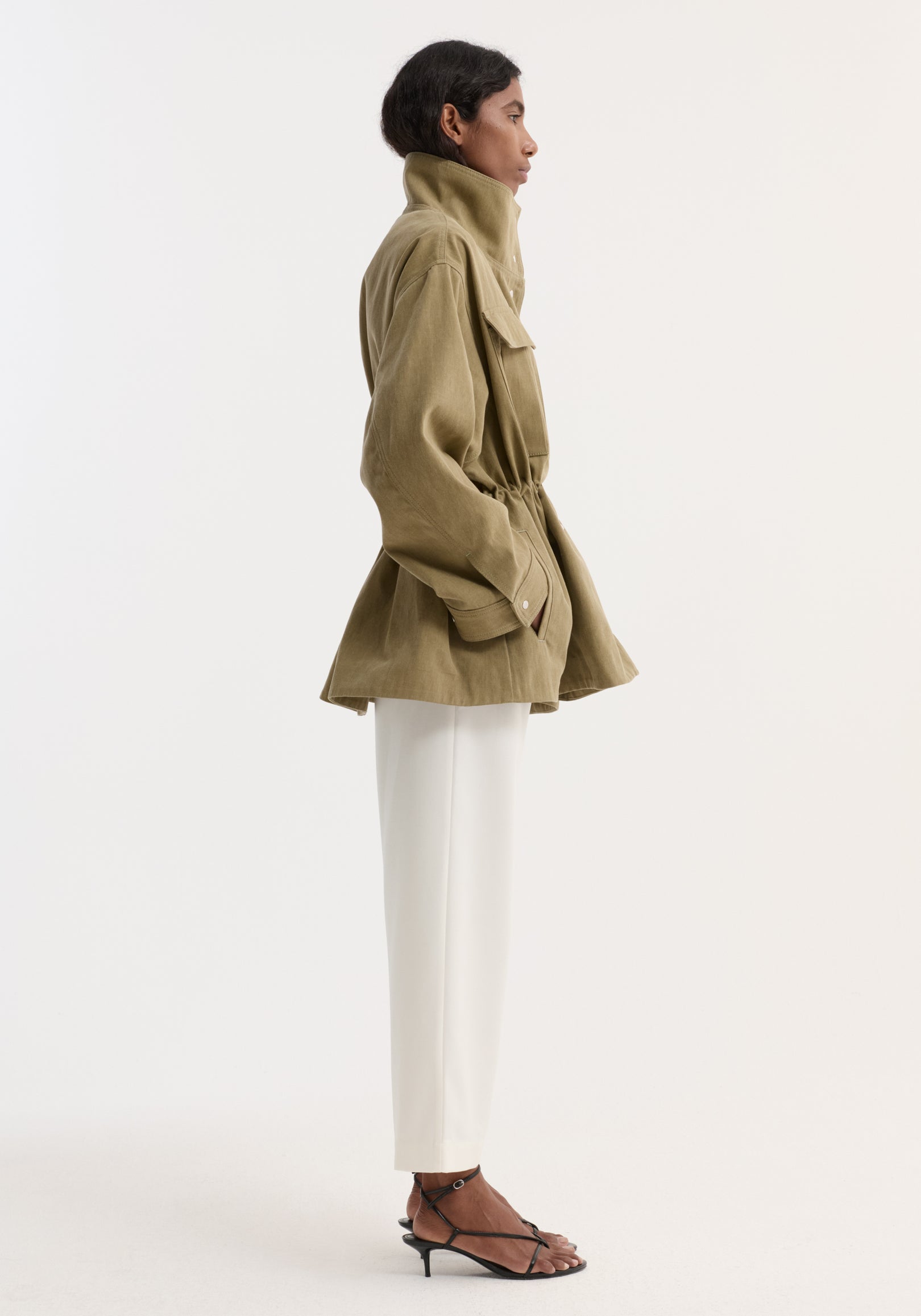 Side profile of a model wearing a sage-colored sculptural safari jacket | sage with a high collar, cinched waist, and flap pockets, paired with white pants and black strappy heels.