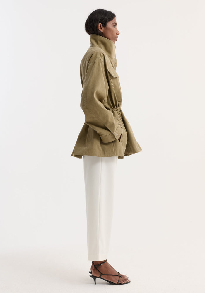 Model wearing Sculptural safari jacket | sage with stand collar, bellows pockets, and elasticated waist, paired with white trousers and black sandals against a plain background.