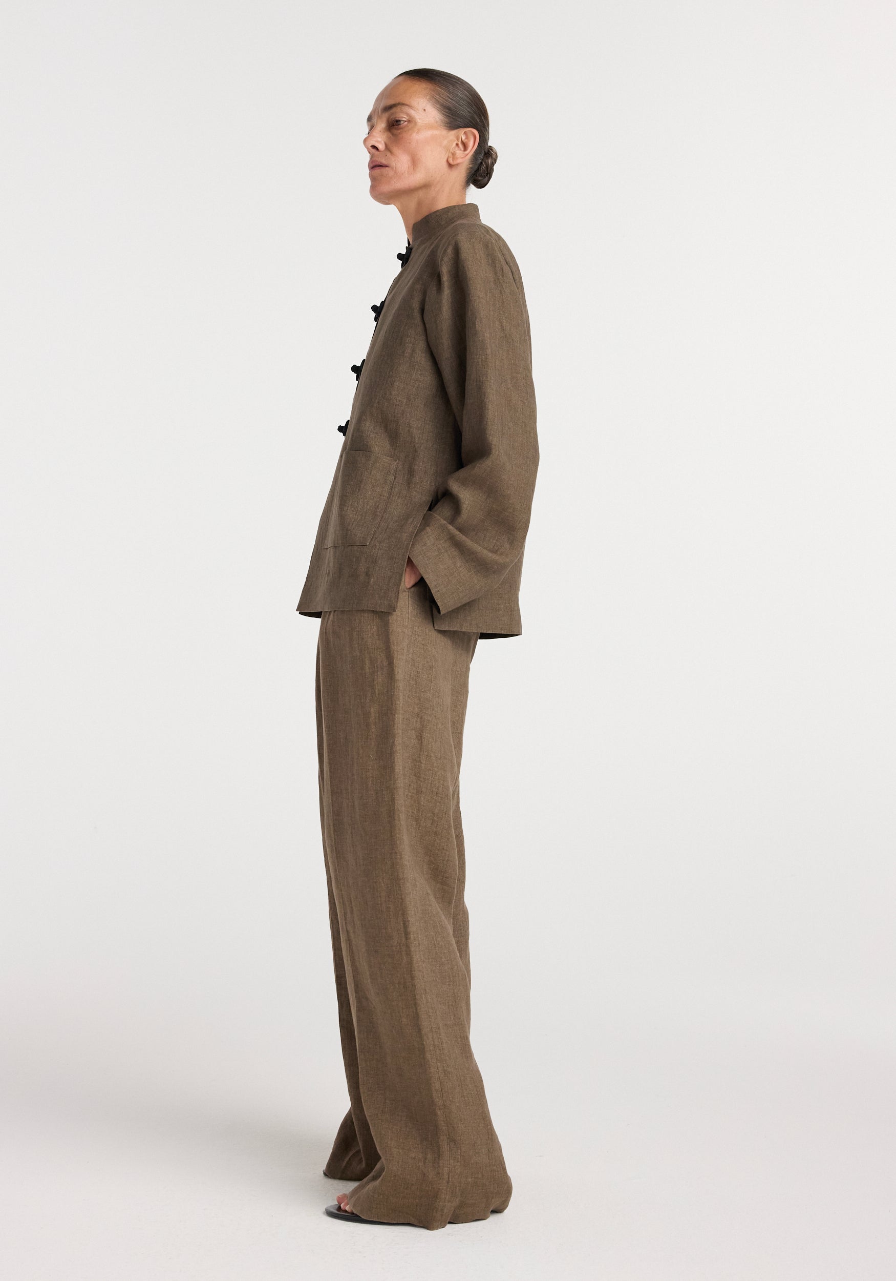 Profile view of a woman wearing a brown melange linen jacket with pankou closures and matching wide-leg linen pants against a plain white background.