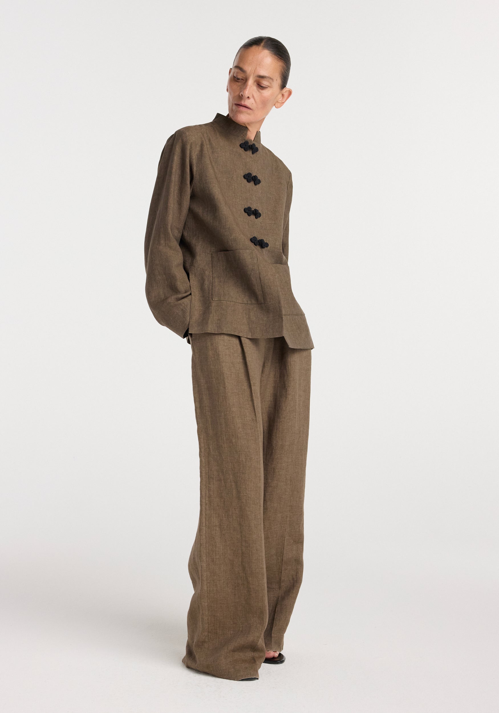 Woman wearing a Pankou closure linen jacket | brown melange with matching wide-leg linen pants, standing against a plain white background looking down.