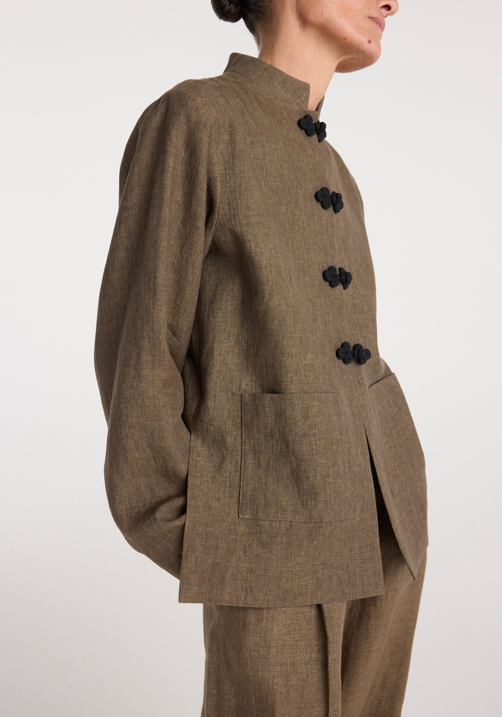 Side view of a person wearing a Pankou closure linen jacket | brown melange featuring a high collar, long sleeves, two front patch pockets, and four black traditional pankou knot closures.