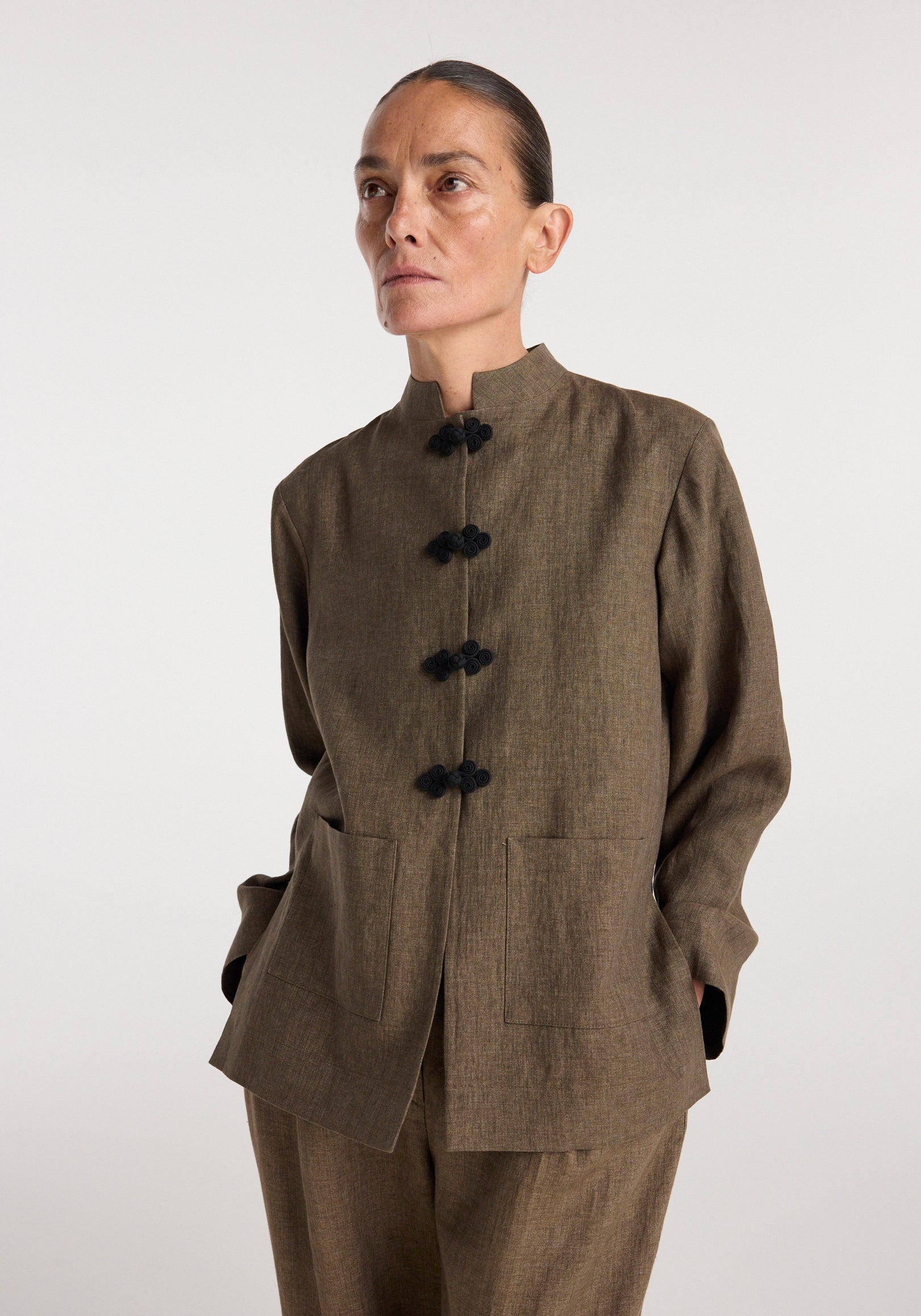 Woman wearing a Pankou closure linen jacket | brown melange with a stand collar and black traditional knot buttons, featuring two front pockets and long sleeves.