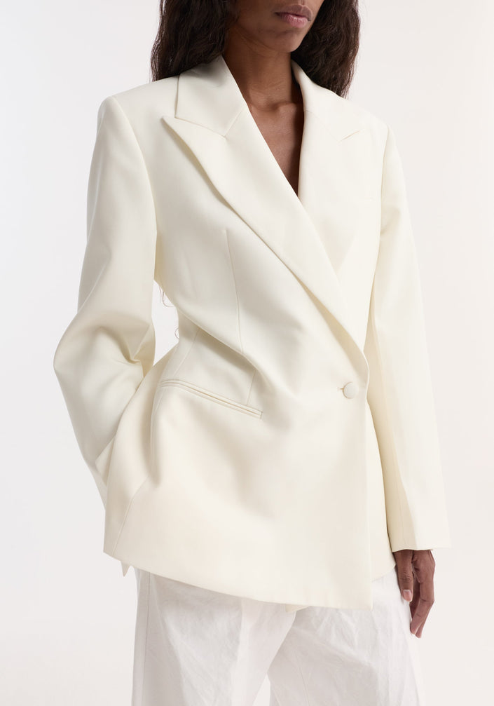 Model in Hourglass shaped blazer, off white, featuring a sharp notch lapel, cinched waist, single button, paired with wide-leg trousers and open-toe sandals.