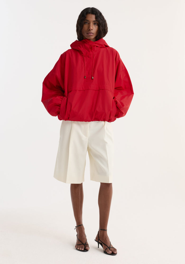 Model wearing Volume silk-blend anorak | red, featuring adjustable hood, front and zip-side pockets, paired with tailored knee-length shorts and black strappy sandals.