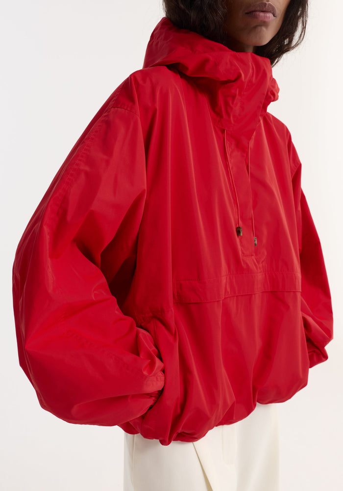 Volume silk-blend anorak in red, featuring a high-collar, adjustable hood, front kangaroo pocket, and elasticized cuffs, worn by model with hands in side pockets.